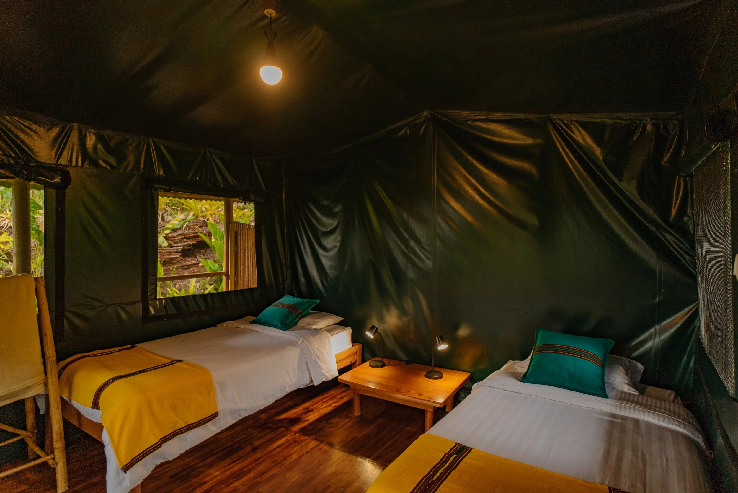 Habitación con paredes de plástico negro, con dos camas con sábanas blancas y mantas amarillas con detalles en negro y rojo. Cada cama tiene una almohada azul con rayas. Hay una mesa de madera con dos lámparas pequeñas encendidas entre las camas y ventanas con cortinas de cañizo que dejan ver vegetación exterior.