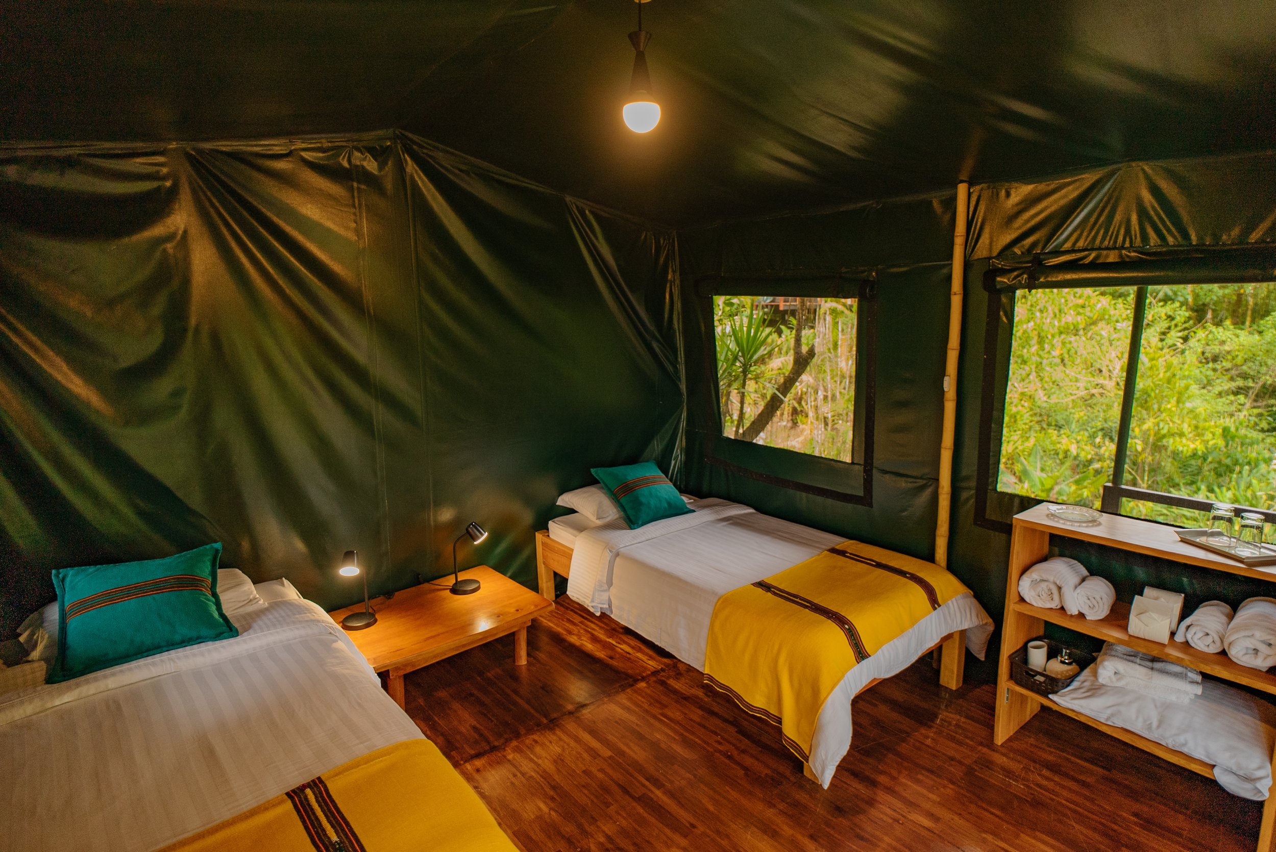 Habitación con tres camas en un espacio con paredes y techo de lona verde, ventanas grandes con vista a la naturaleza, y muebles de madera, incluyendo mesas y estantes, con ropa de cama en colores blanco, amarillo y verde.