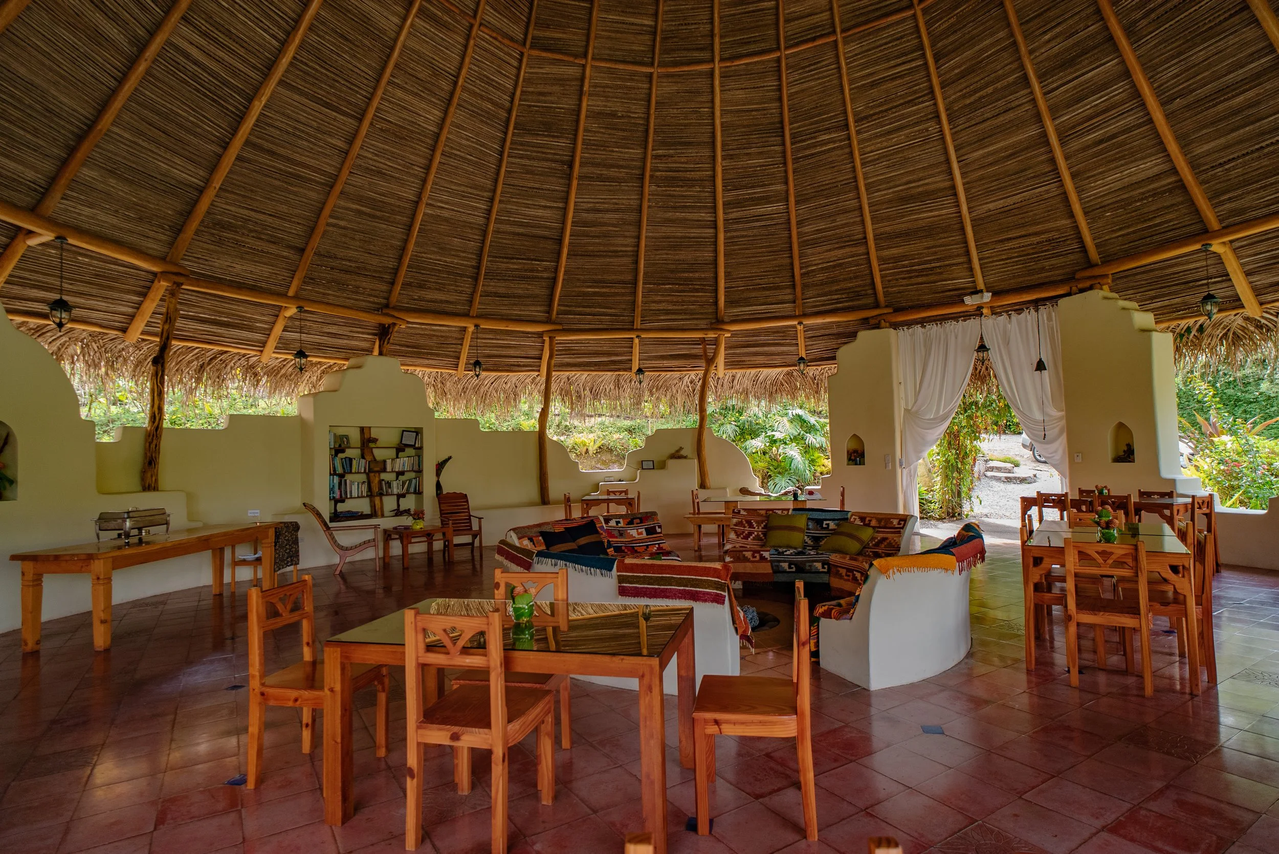Sala de estar con techo de palma, paredes encaladas, paredes con nichos, muebles de madera, sillones con textiles coloridos y cestas, con vista a un jardín exterior con plantas verdes