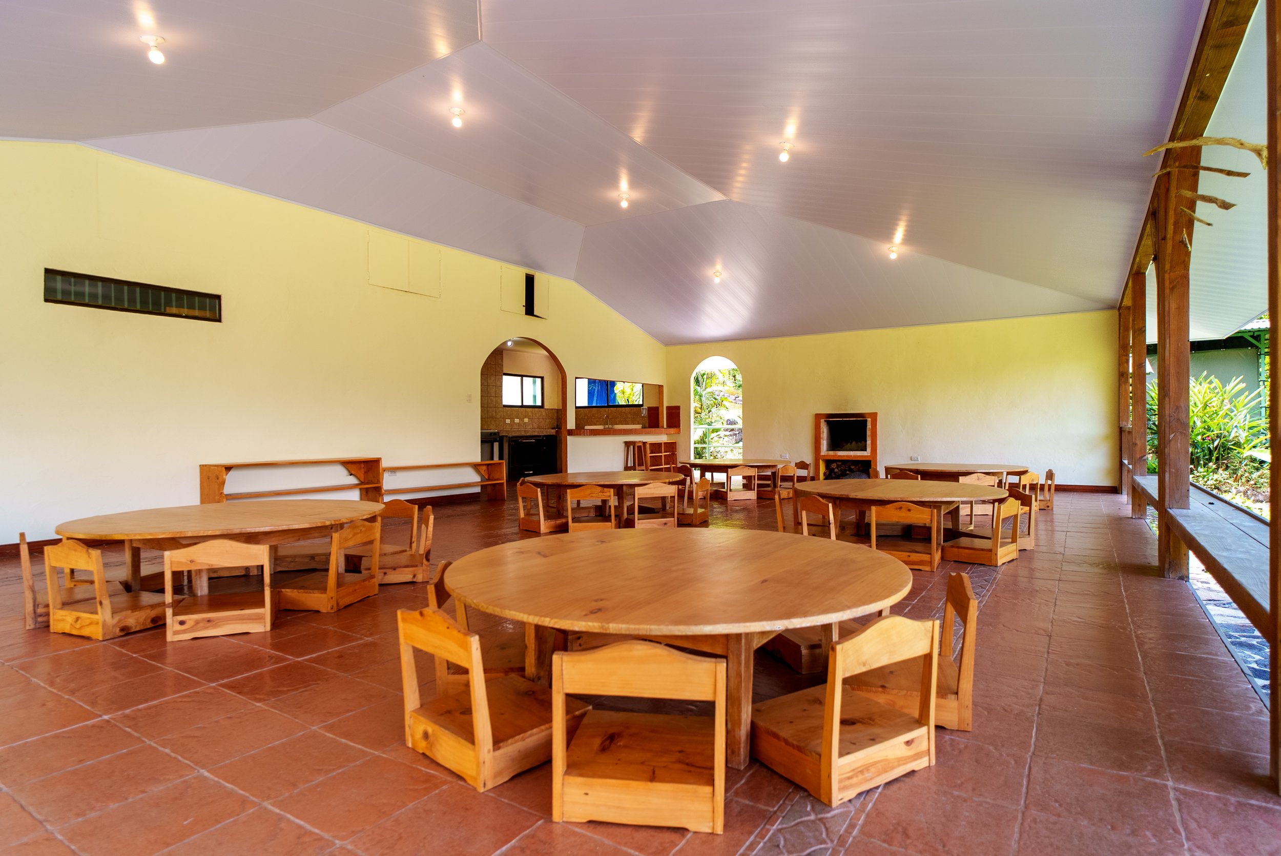 Interior de un espacio amplio con mesas redondas y sillas de madera. Paredes de color amarillo y un techo beige con luces. Ventanas y puertas que muestran vegetación exterior.