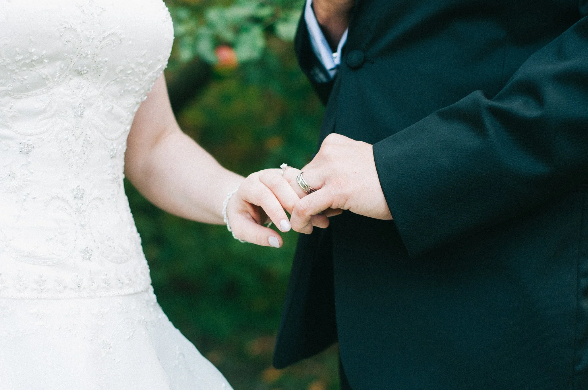 Close-up of a bride and groom holding hands, showing wedding rings, with a blurred green background.