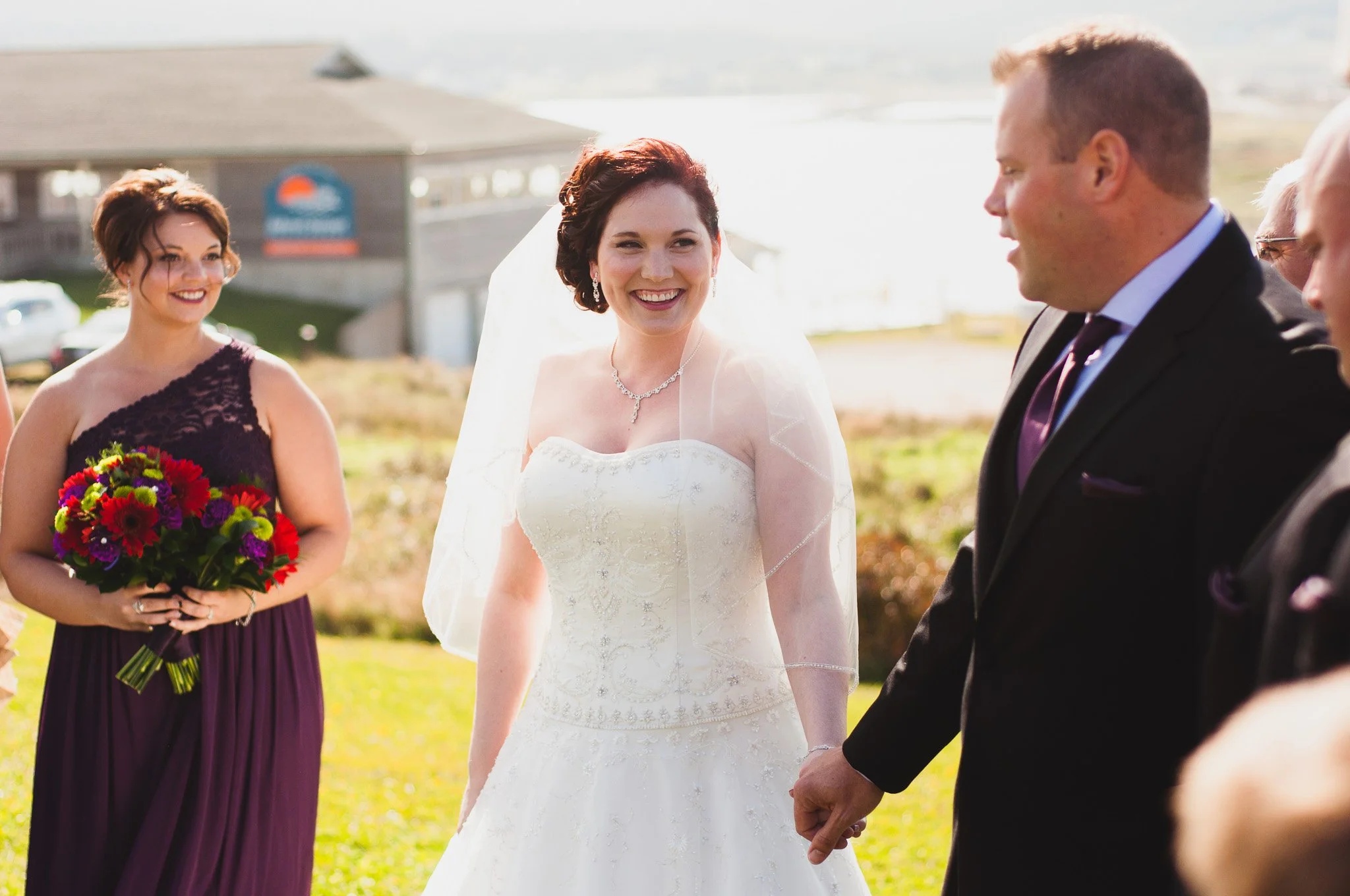 Bride and groom holding hands during outdoor wedding ceremony with bridesmaid holding a bouquet, bright sunlight, and a building in the background.