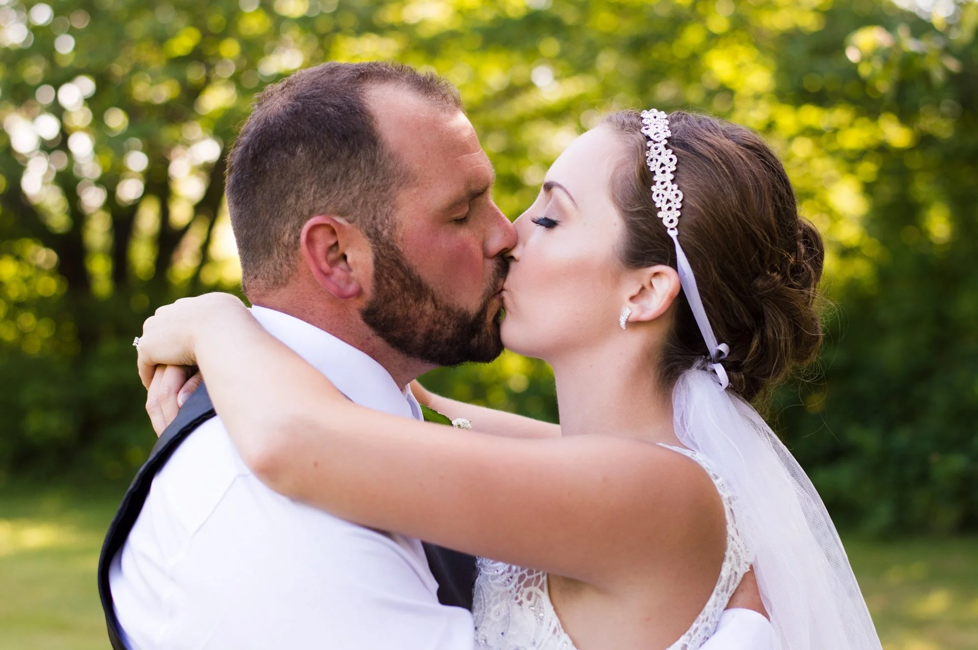 A newlywed couple sharing a kiss outdoors, surrounded by green trees.