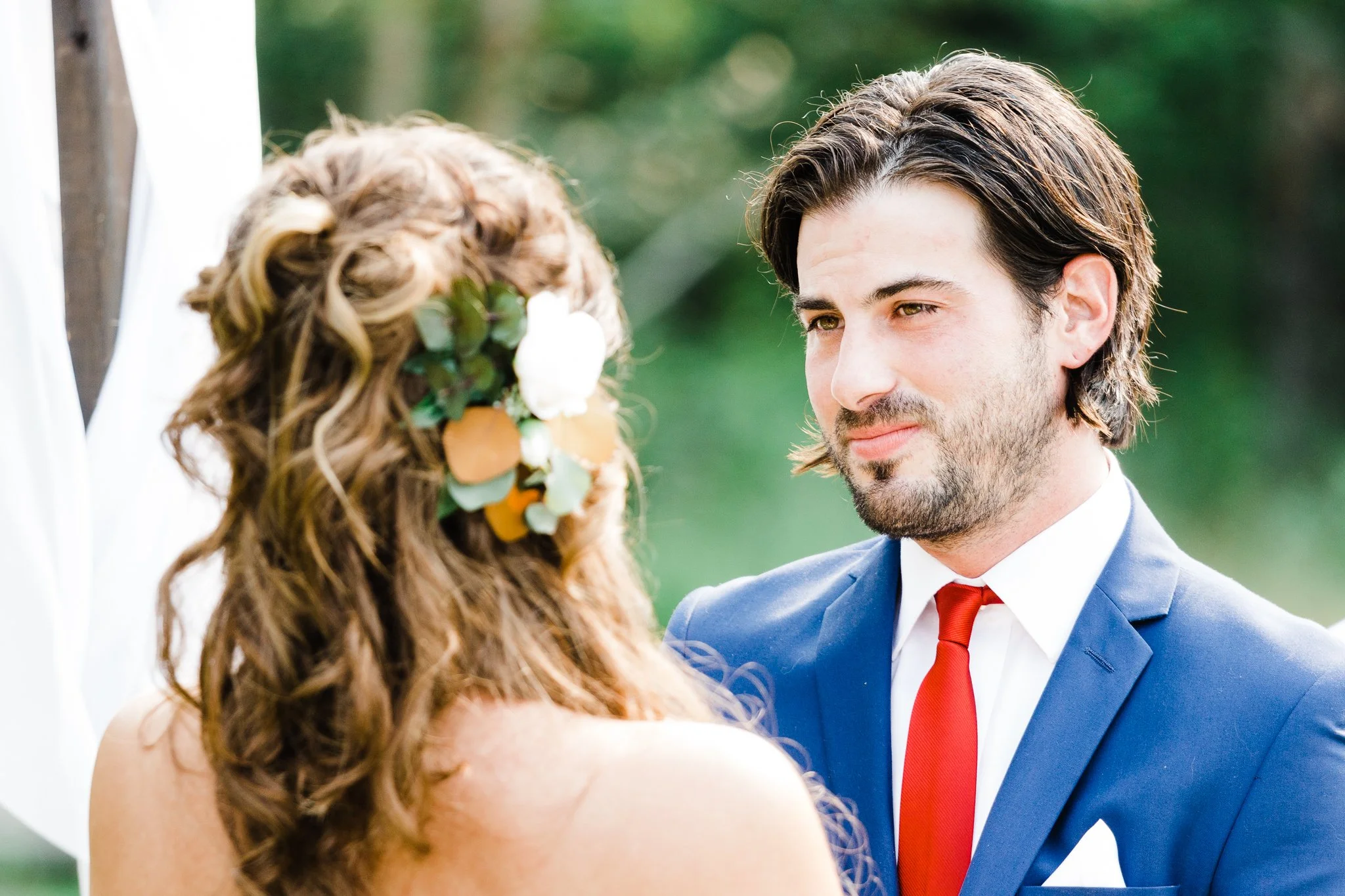 A man and woman exchanging vows during their wedding ceremony outdoors, with the woman wearing a floral hairpiece and the man in a blue suit with a red tie.