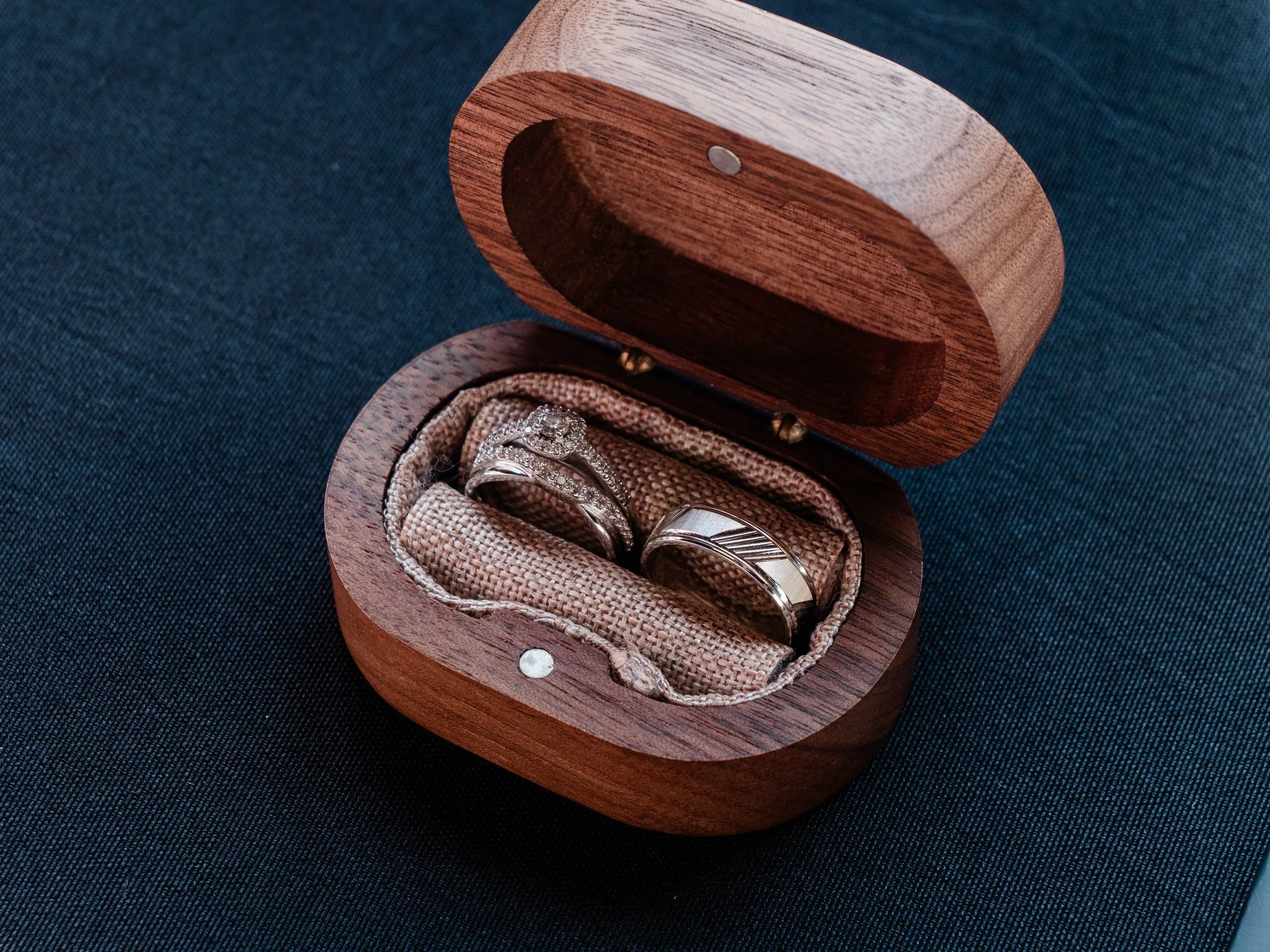 Open wooden jewelry box containing two wedding rings, one with a diamond and the other plain, resting on a brown fabric inside the box.