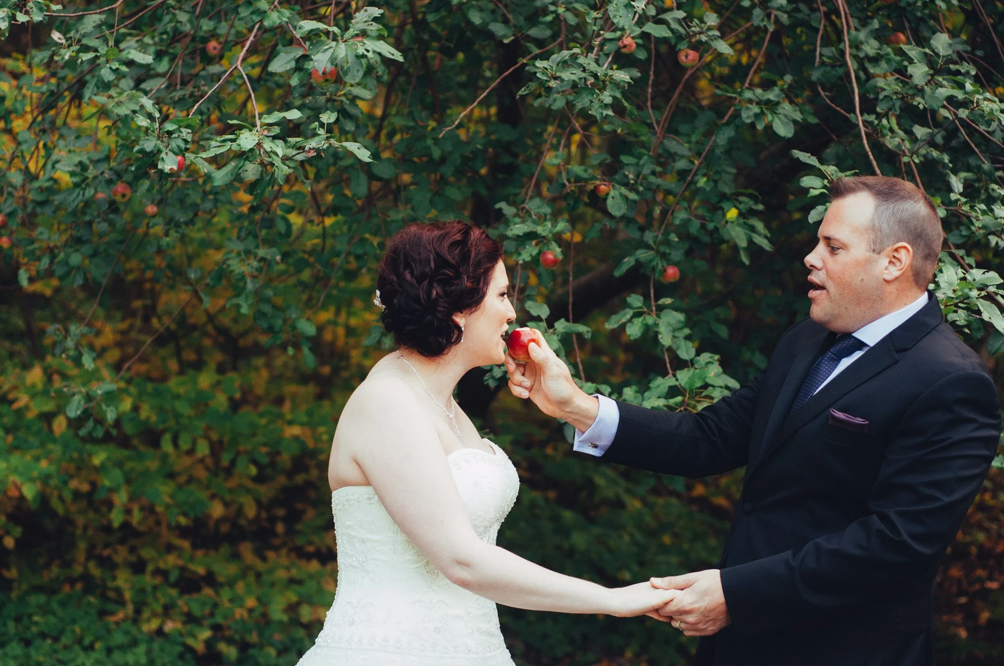 A bride and groom holding hands outdoors with an apple orchard background. The groom is feeding the bride an apple.