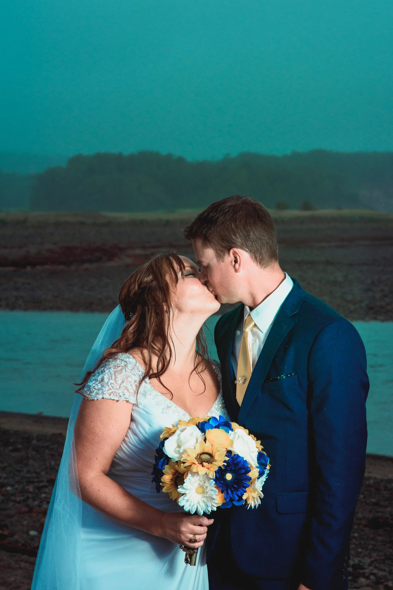 A bride and groom sharing a kiss on their wedding day outdoors near water, with a landscape of trees and hills in the background.