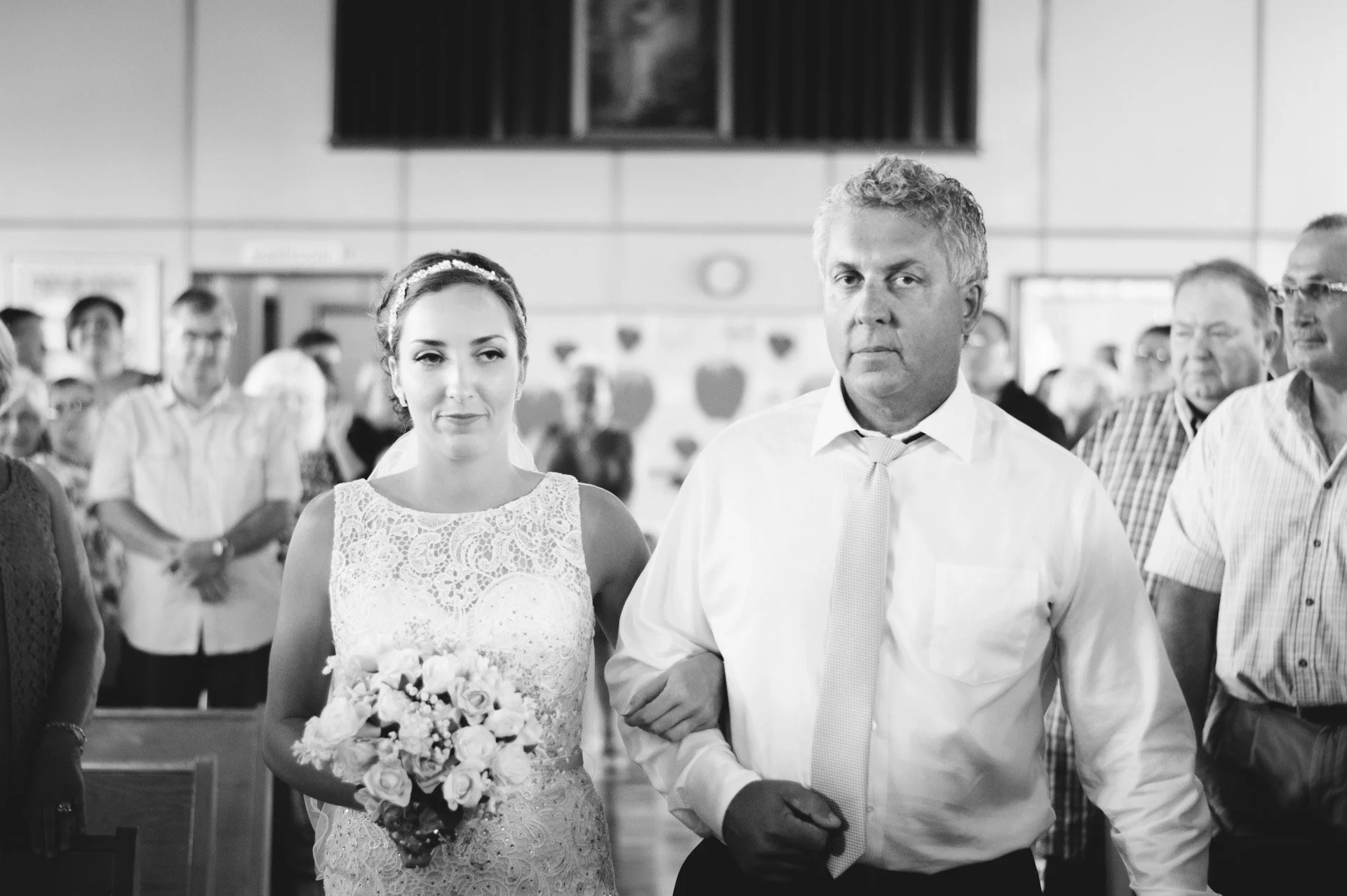 A bride in a lace wedding dress holding a bouquet walking down the aisle with a man, likely her father, at a wedding ceremony in a church filled with guests.