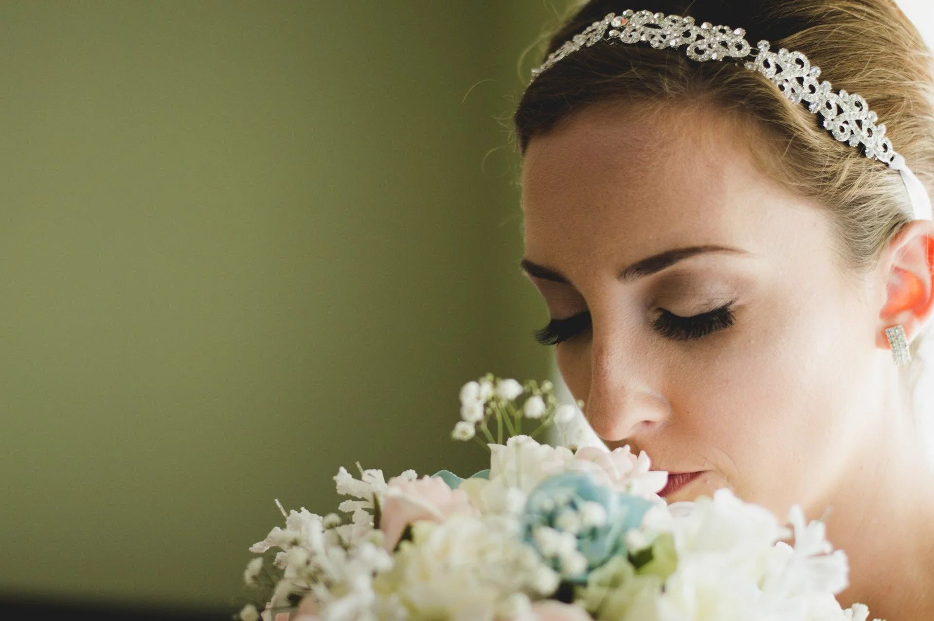 Close-up of a bride with a diamond headband and earrings, holding a bouquet of white and pastel flowers, with eyes closed and a serene expression.