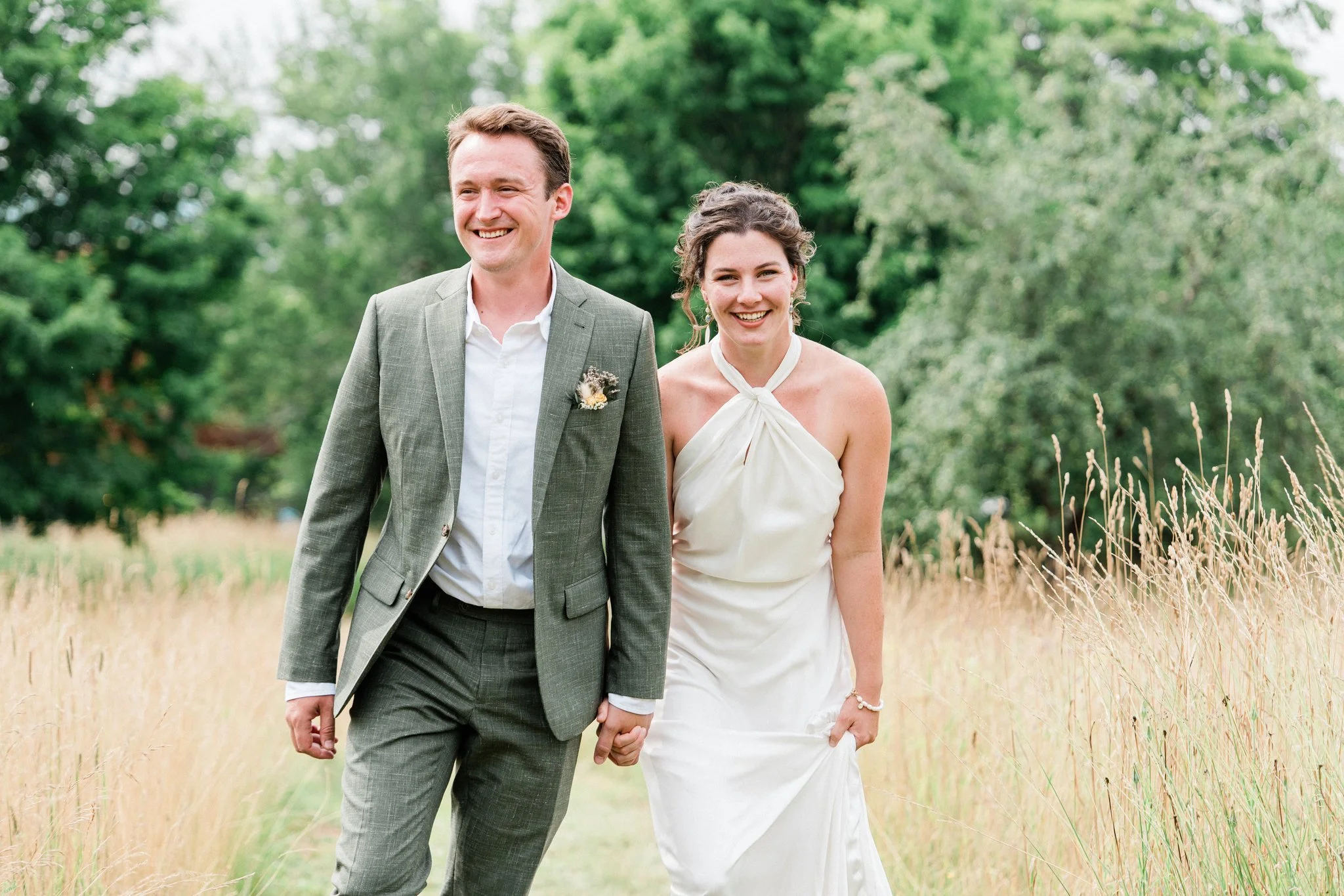 A smiling couple, dressed in wedding attire, walking hand in hand through a grassy field with trees in the background.