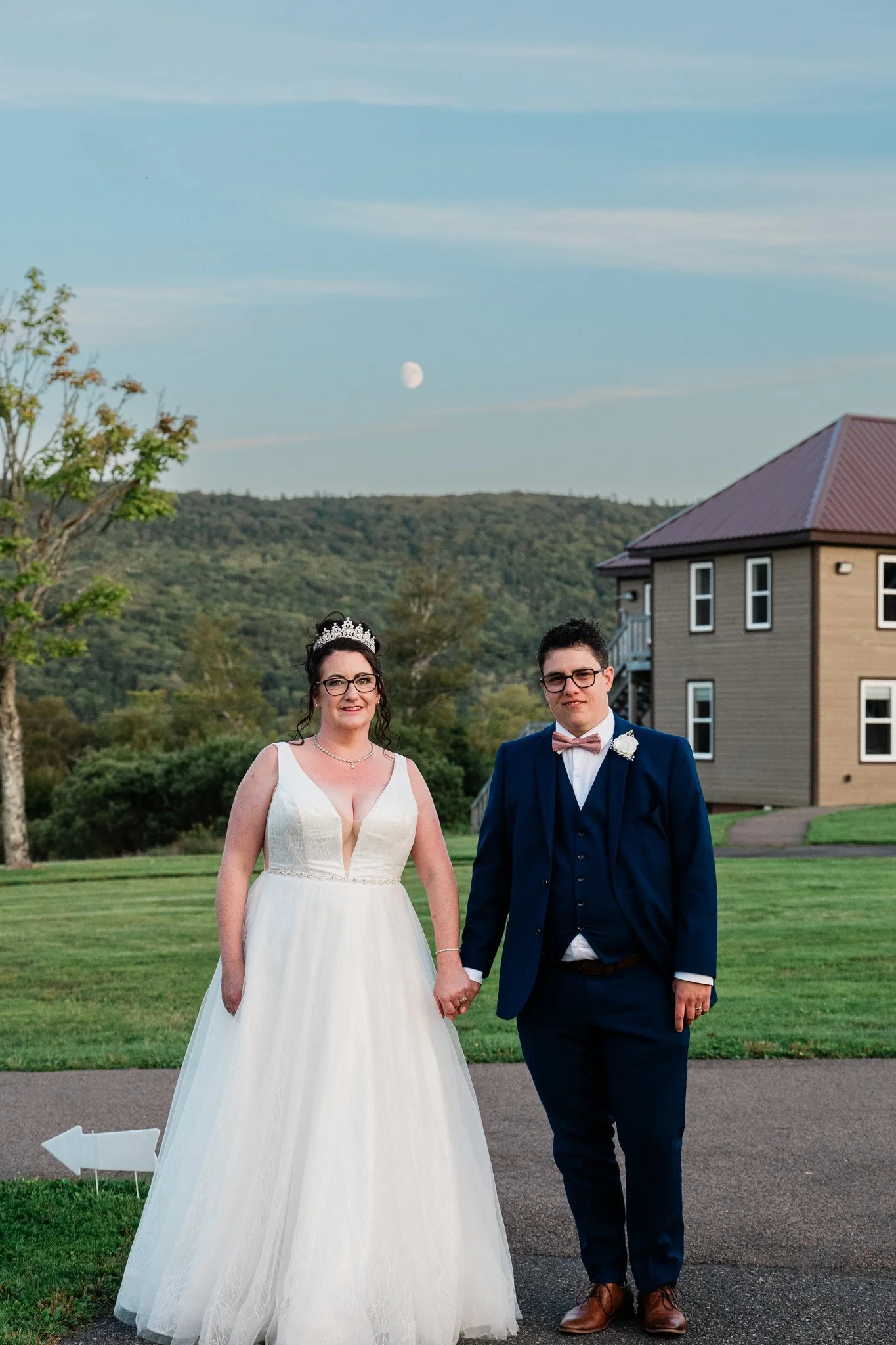 A bride and groom holding hands outdoors during their wedding, with a house and mountain in the background under a partly cloudy sky and visible moon.