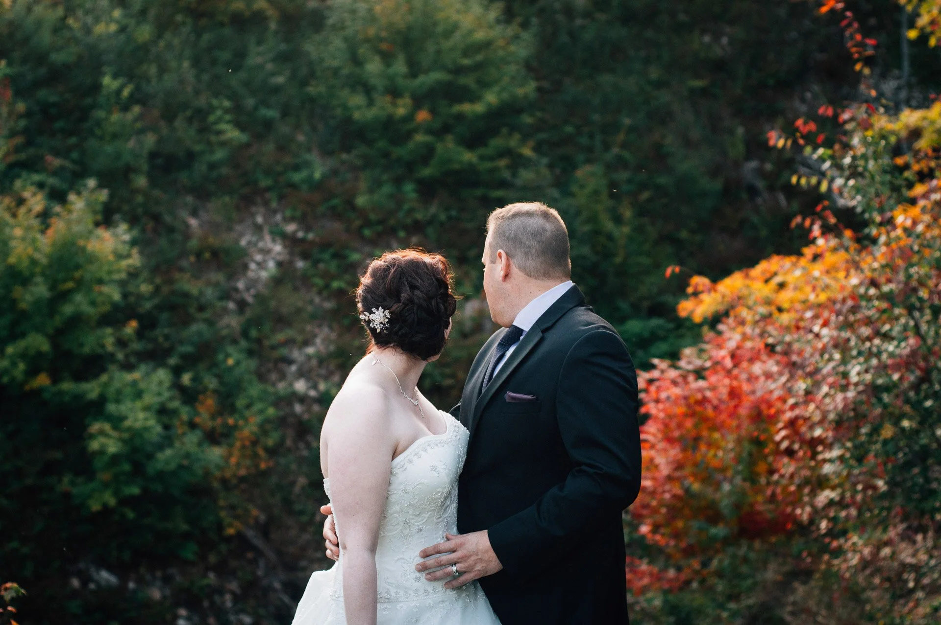 A bride and groom stand closely together outdoors during autumn, with colorful fall foliage in the background.