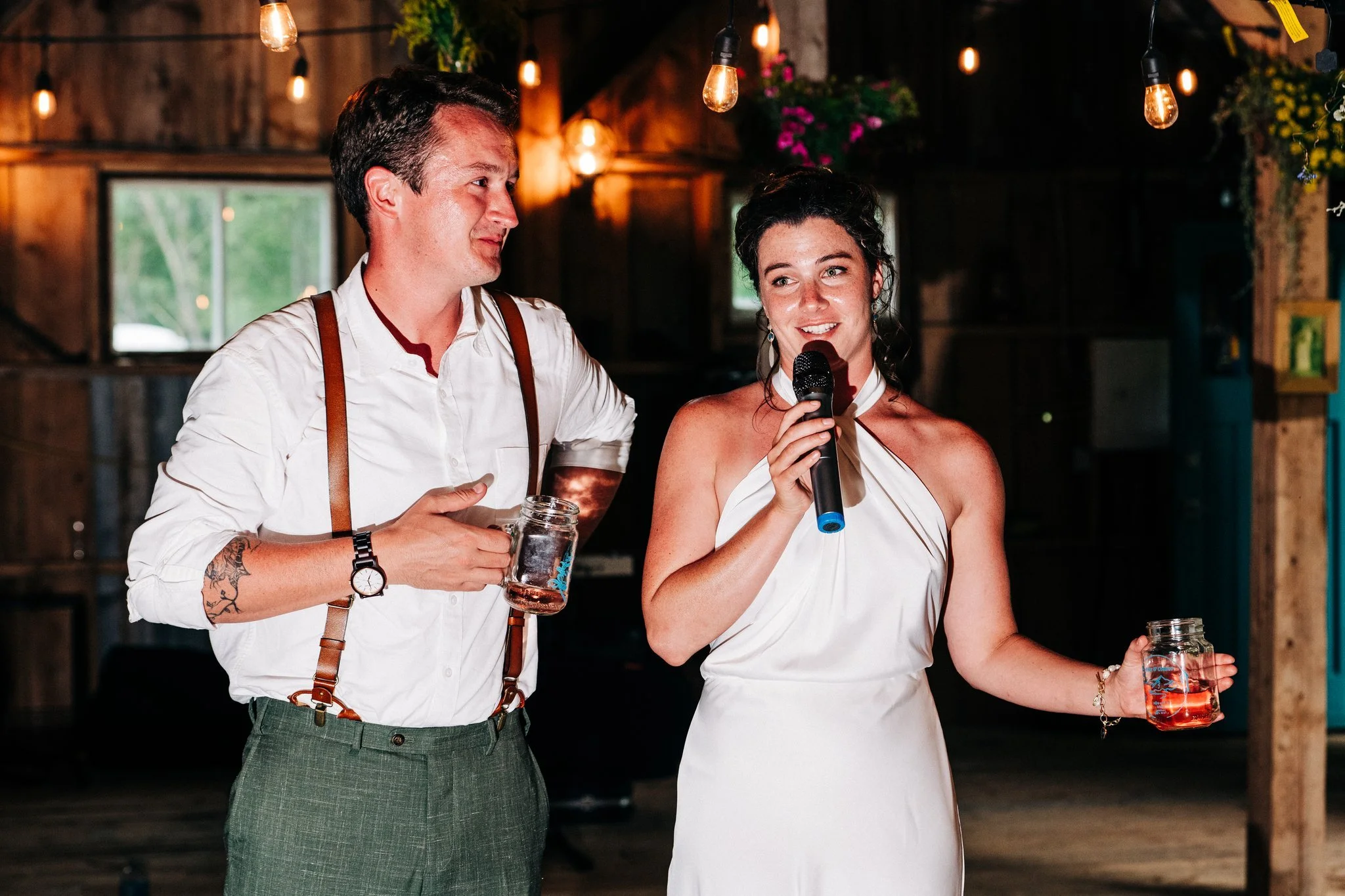 A man and a woman at a rustic indoor event, with the woman speaking into a microphone. The man holds a drink in a jar. Both are dressed formally, with the woman in a white dress and the man in a white shirt with suspenders. Hanging lights and flowers
