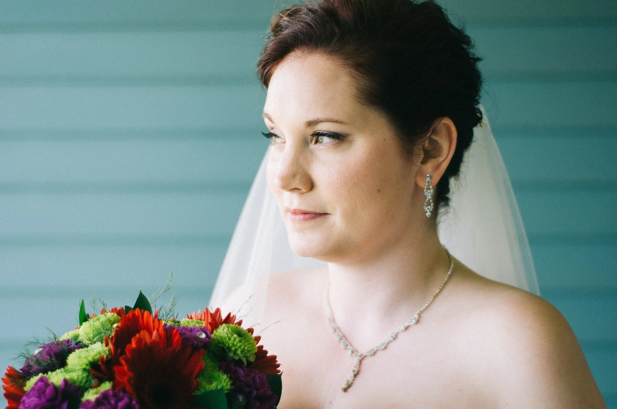 A bride with short dark hair, wearing earrings and a necklace, holding a colorful bouquet of red, purple, and green flowers, and looking to the side with a soft expression.