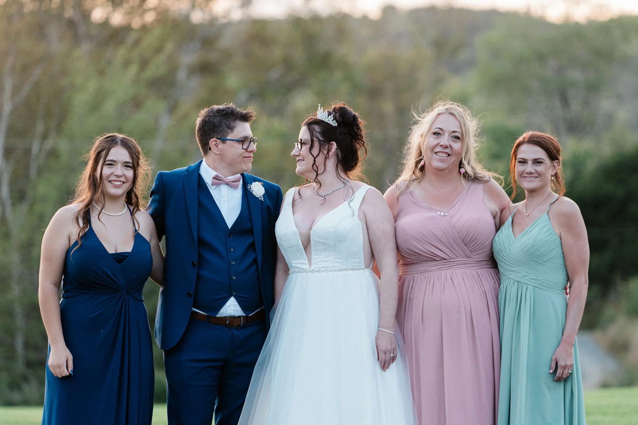A group of five women and one man dressed in formal attire, standing outdoors during a wedding, with trees in the background.