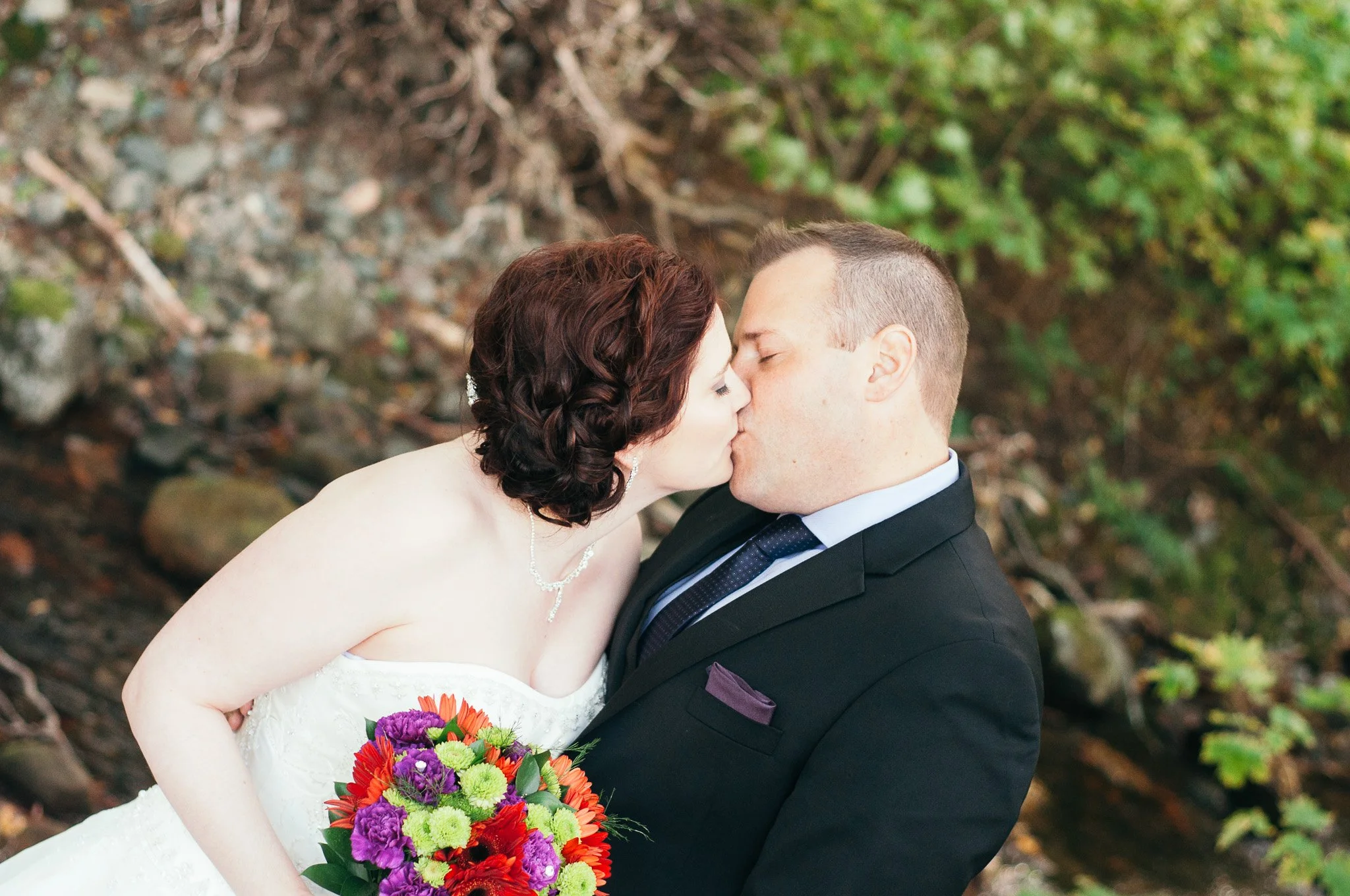 Bride and groom kissing outdoors, bride holding colorful bouquet, trees and rocks in background.