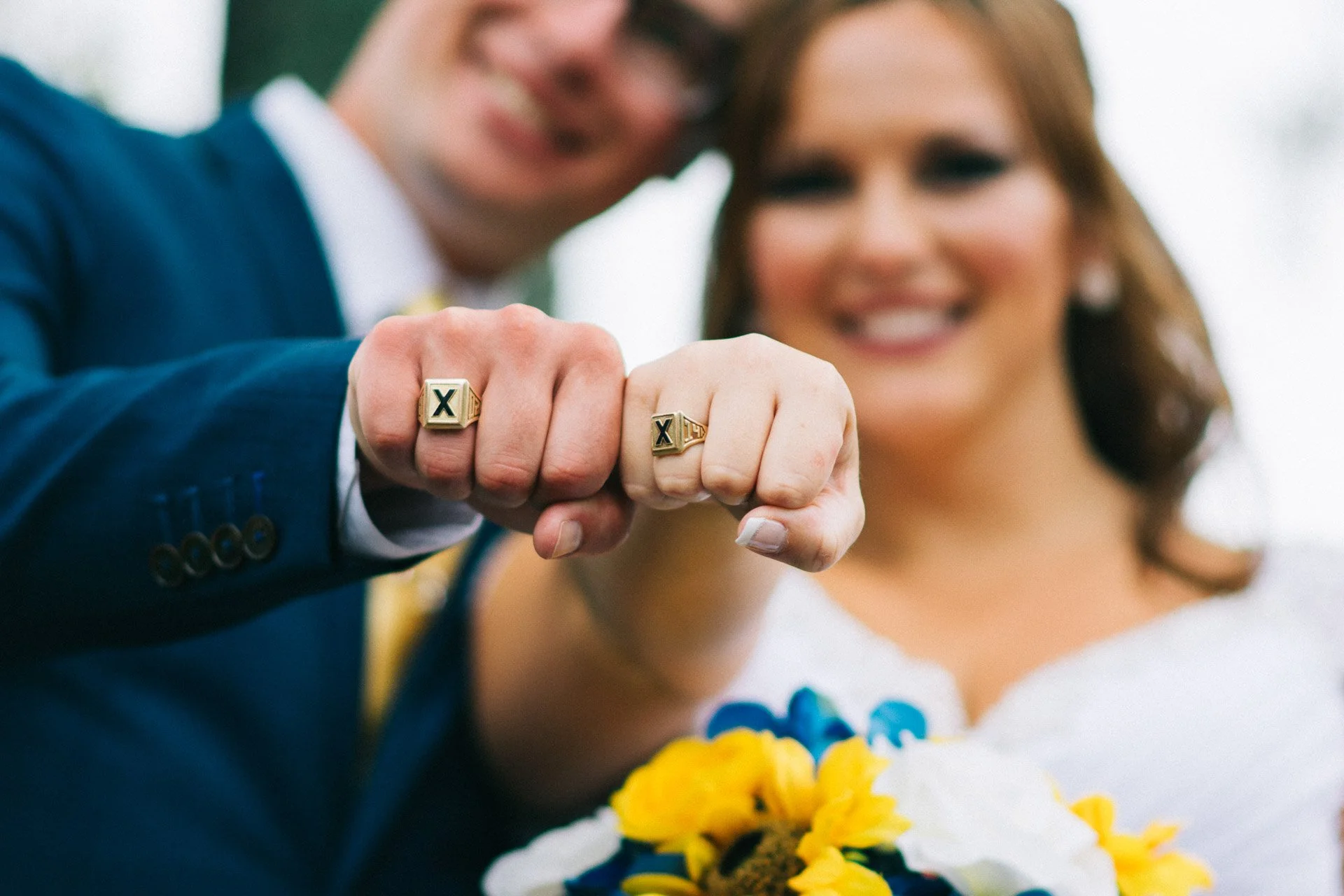 Close-up of a couple’s fists with matching gold rings, with the woman holding a bouquet of yellow and blue flowers in the background.