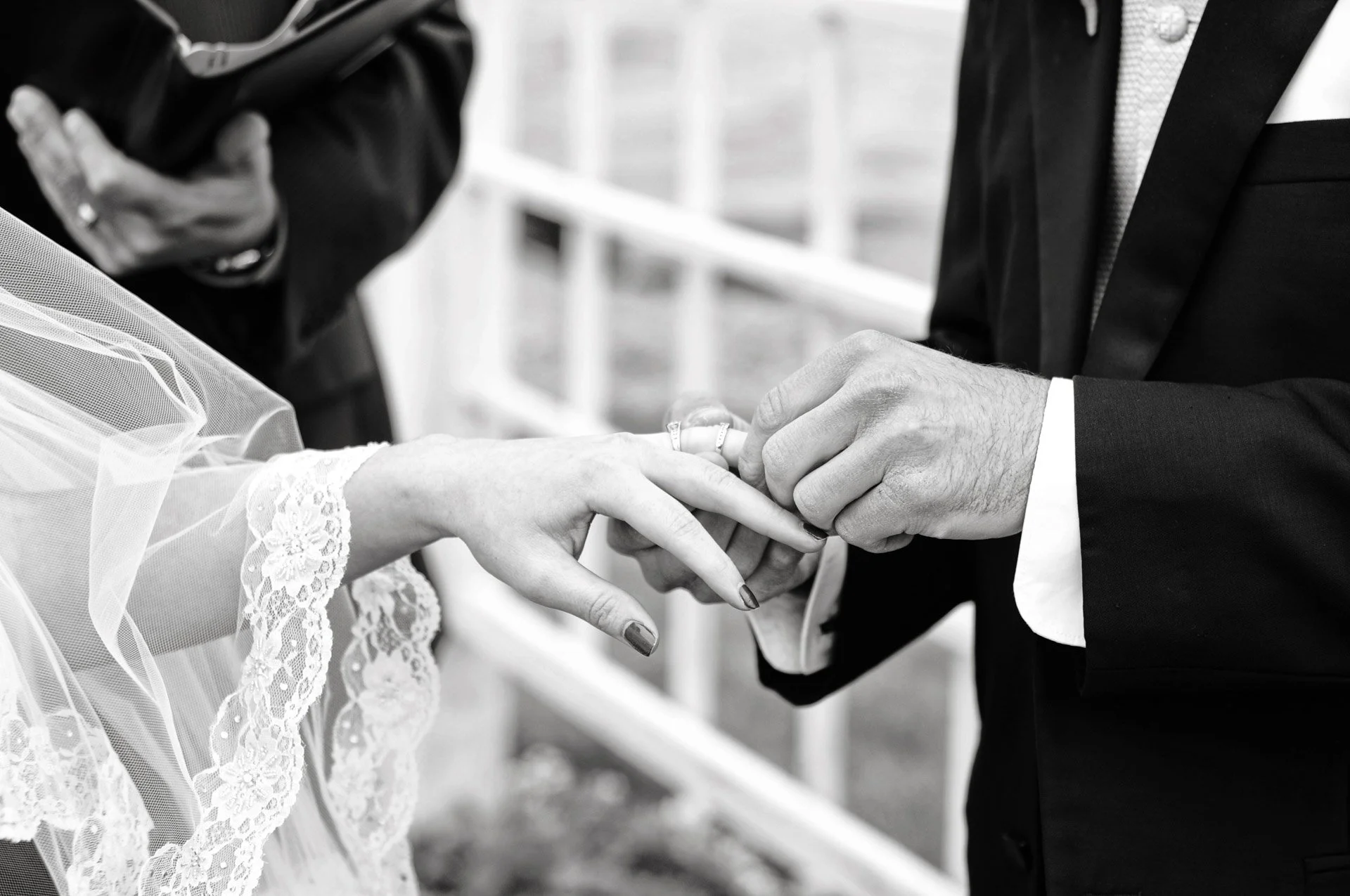 A black and white photo of a bride and groom exchanging rings during a wedding ceremony, with the bride's hand extended and the groom placing a ring on her finger.