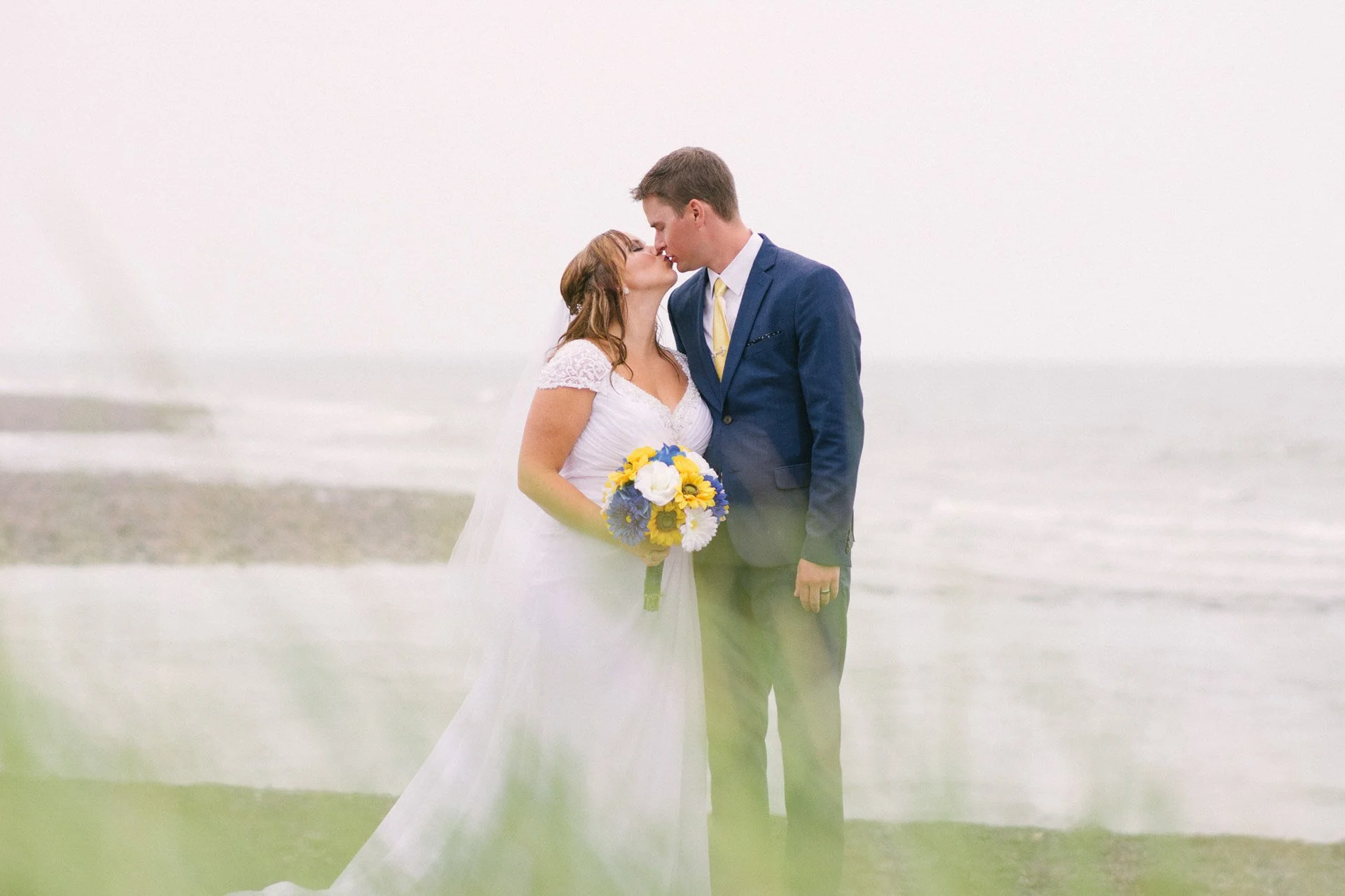 A bride and groom standing on a beach, kissing, with the ocean in the background. The bride is holding a bouquet of yellow, white, and blue flowers.