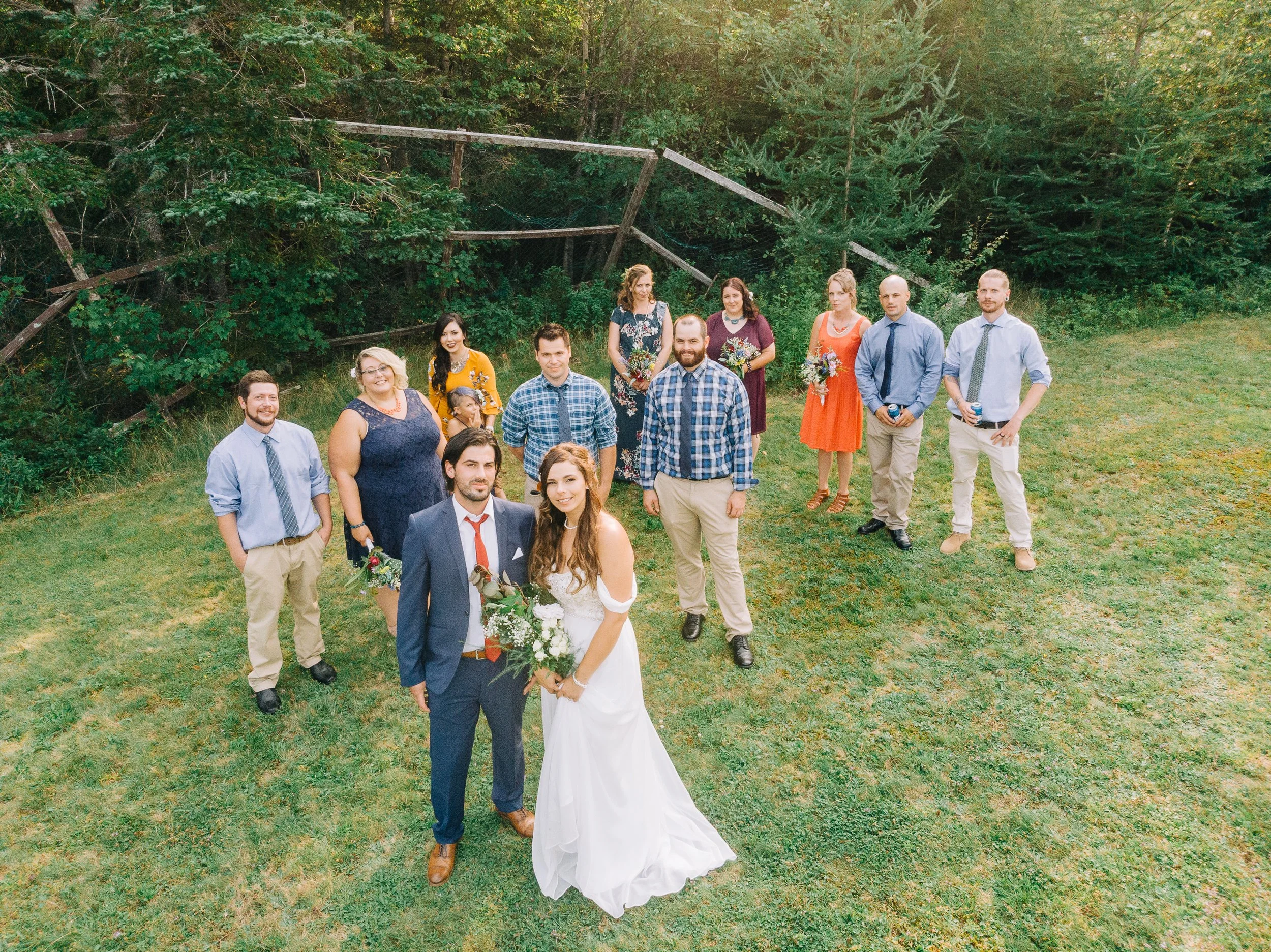 Wedding group photo outdoors with the bride and groom in front, surrounded by family and friends on a grassy field with trees in the background.