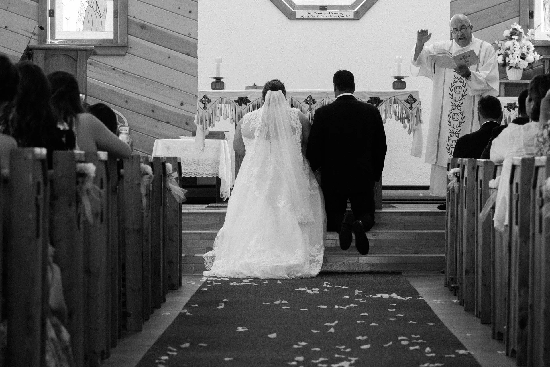 Bride and groom kneeling at altar during wedding ceremony in a church, officiated by a priest, with guests seated on wooden pews. Black and white photo.