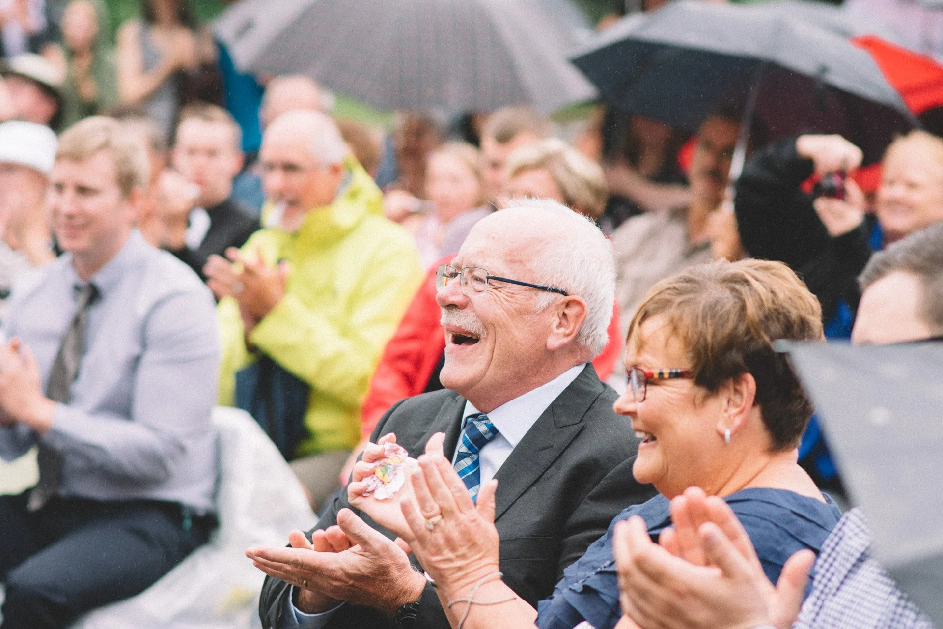A group of people sitting outdoors, holding umbrellas, and smiling or laughing, during a possibly rainy event.