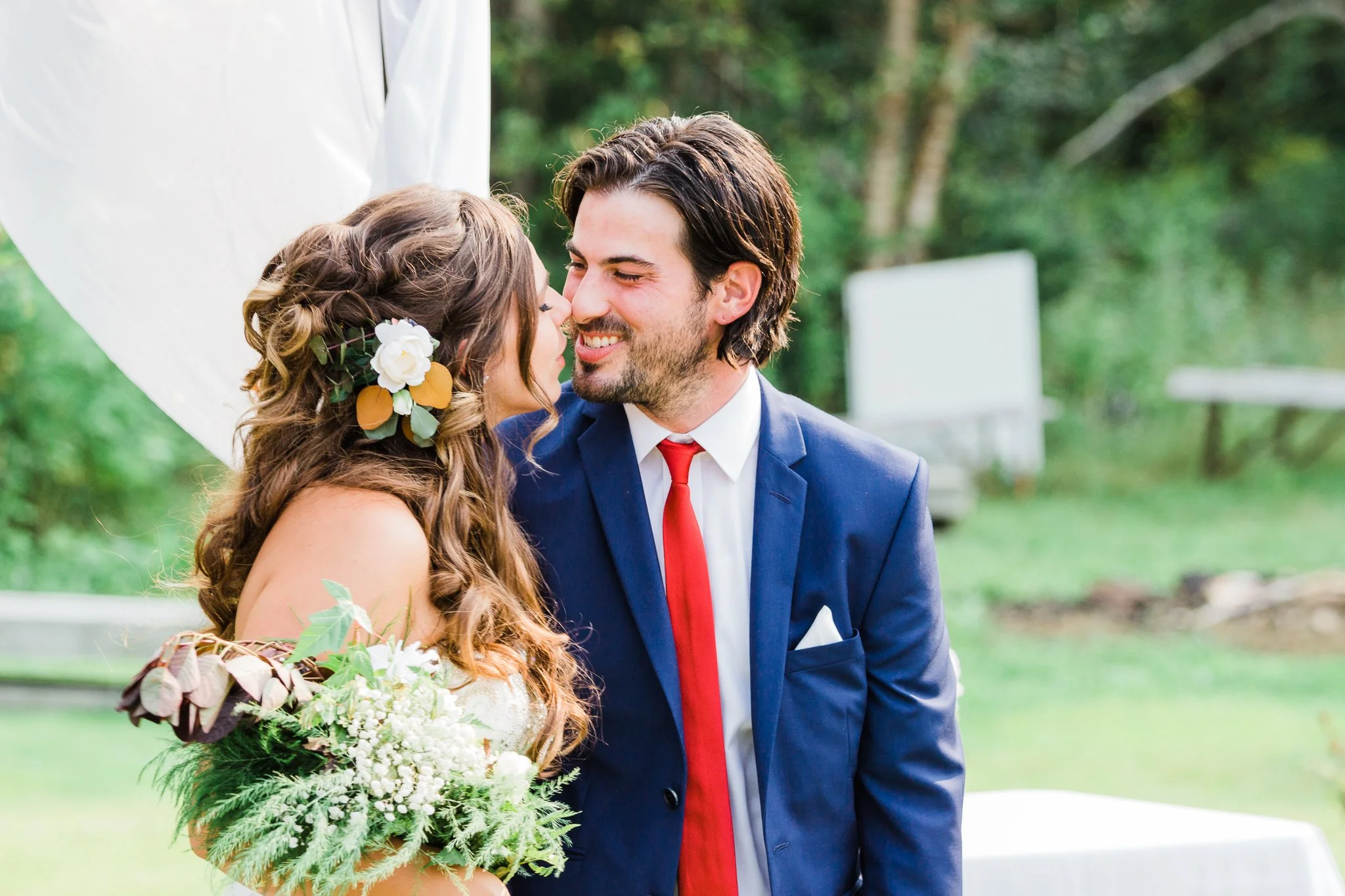 A bride and groom with noses touching, smiling, outdoors with trees and a white backdrop in the background.