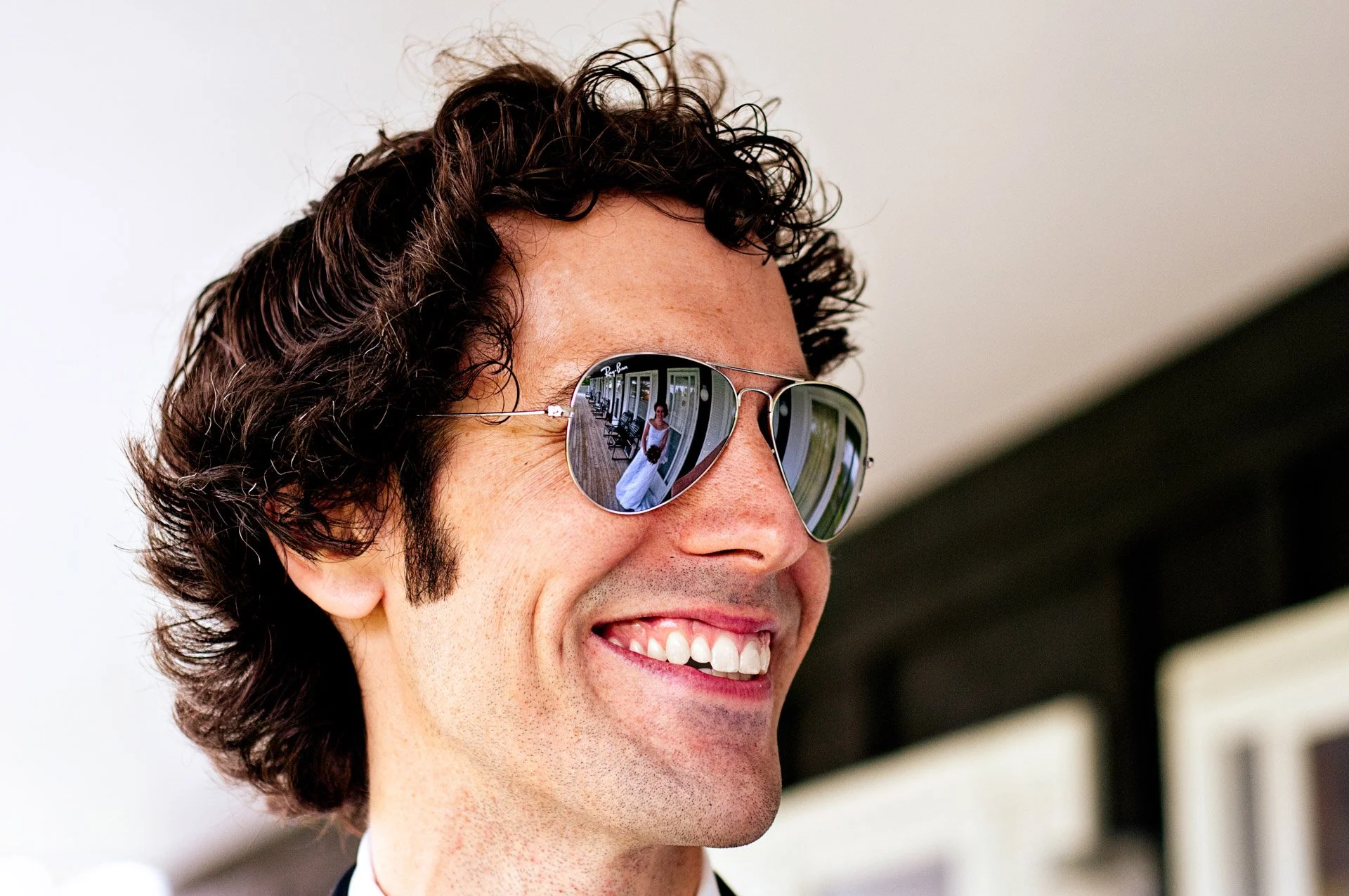 A man with curly dark hair and a wide smile wearing reflective sunglasses, showing a woman in a white dress reflected in his sunglasses.