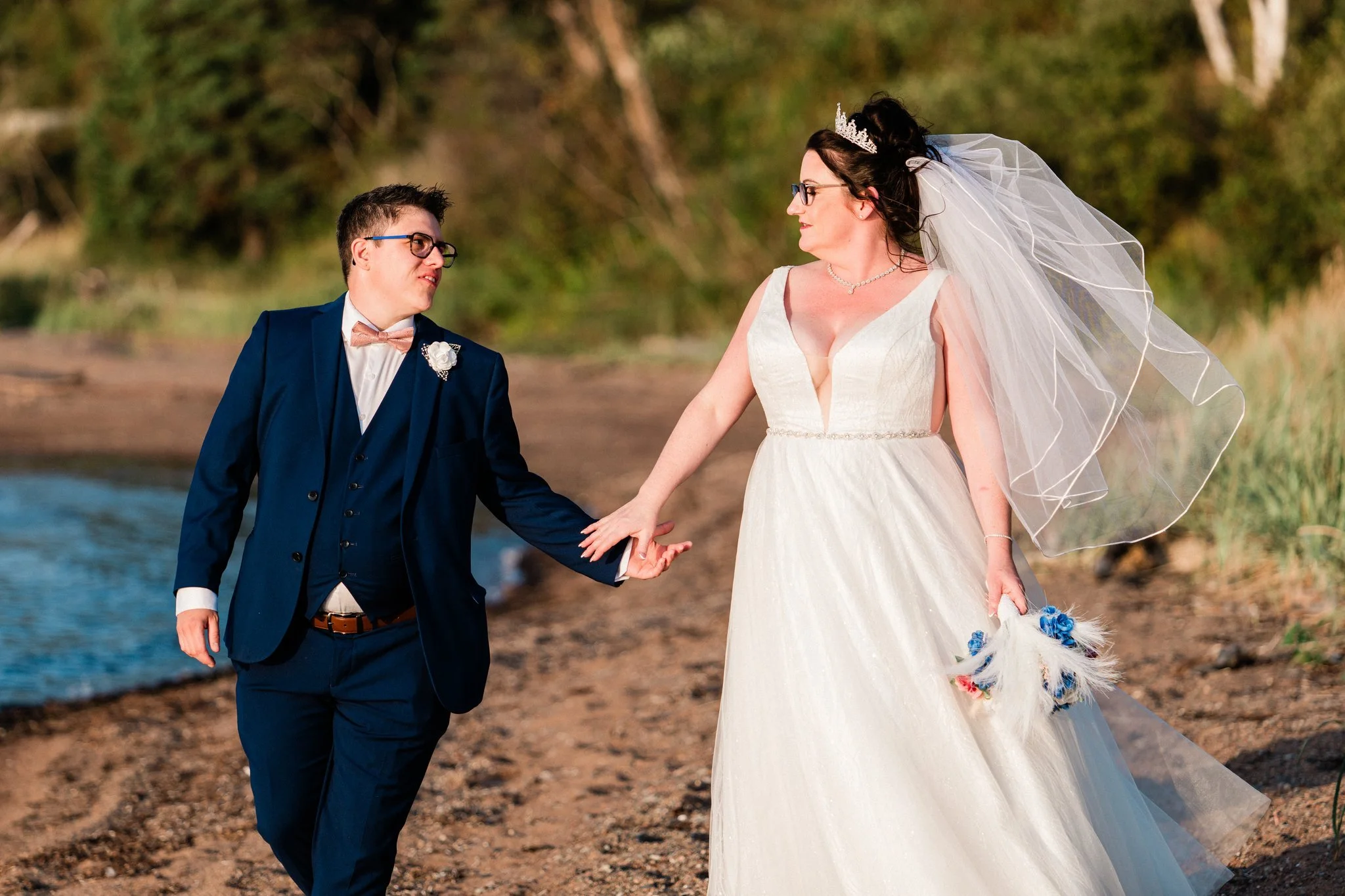 A bride and groom holding hands on a beach, with the bride wearing a white wedding dress and veil, and the groom in a navy suit with bow tie, both looking at each other.