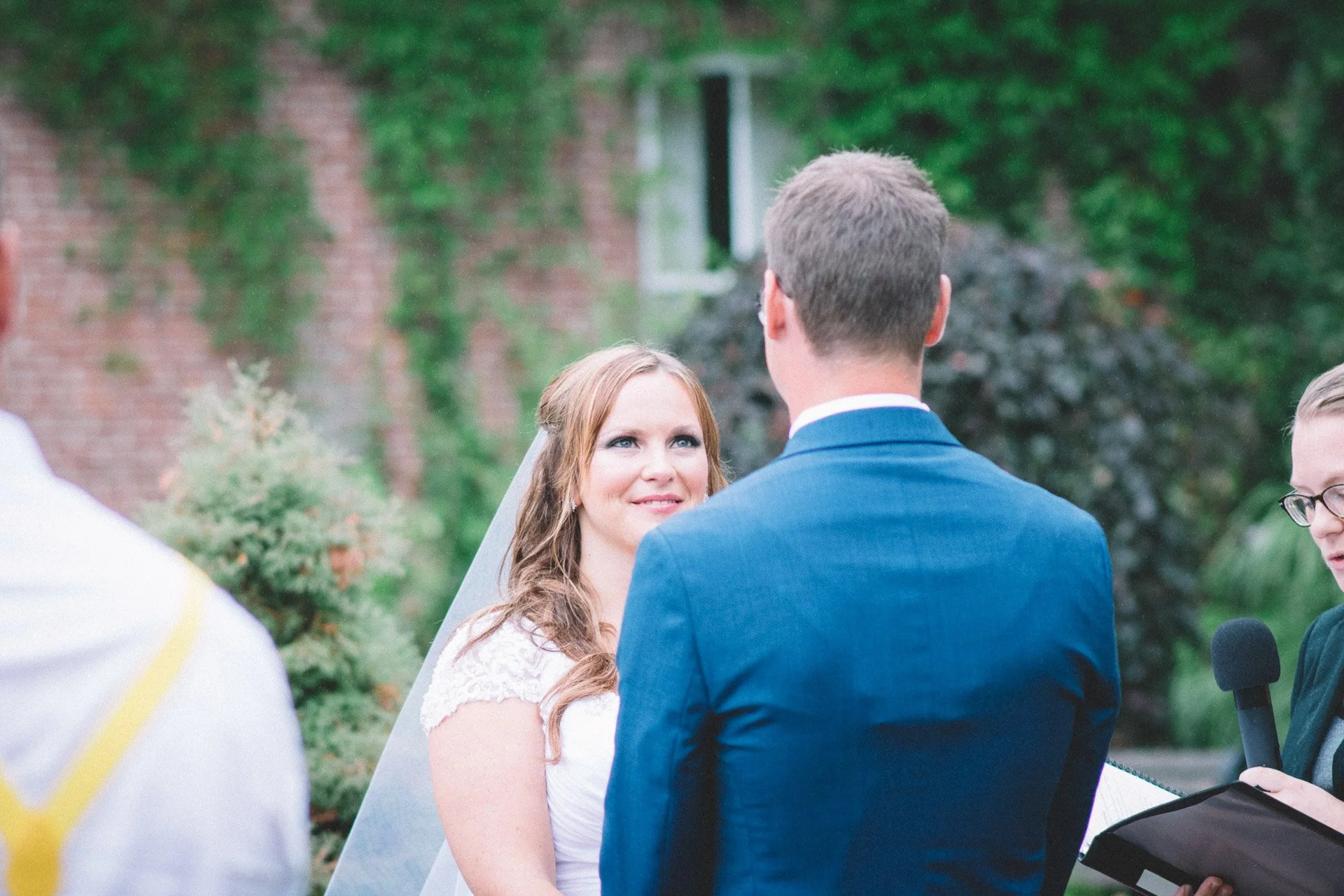 A bride and groom exchange vows during their outdoor wedding ceremony. The bride is looking at the groom while speaking. An officiant holding a microphone and a book is visible on the right. Greenery and brick wall in the background.