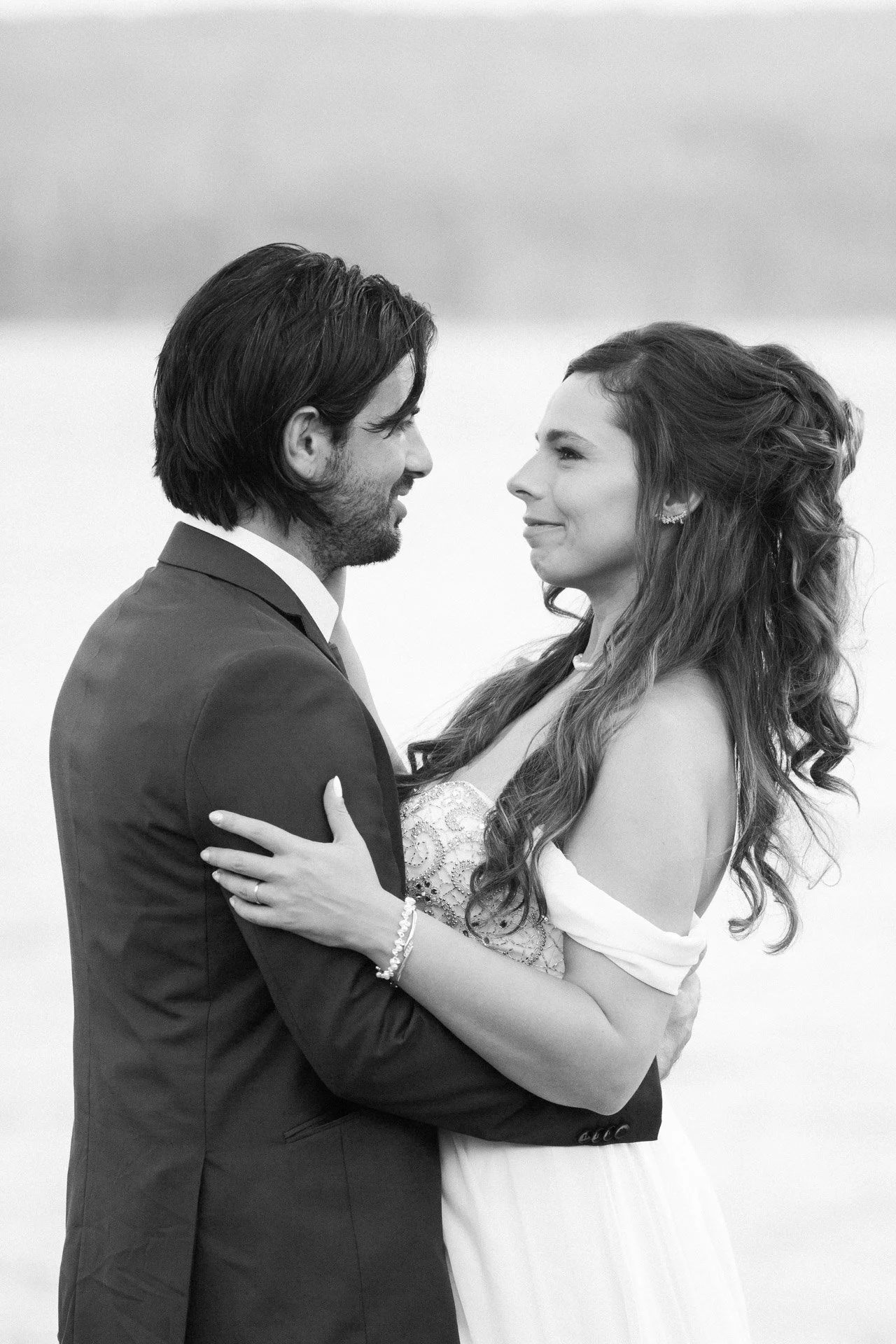 A black and white photo of a couple holding each other, looking into each other's eyes, outdoors.