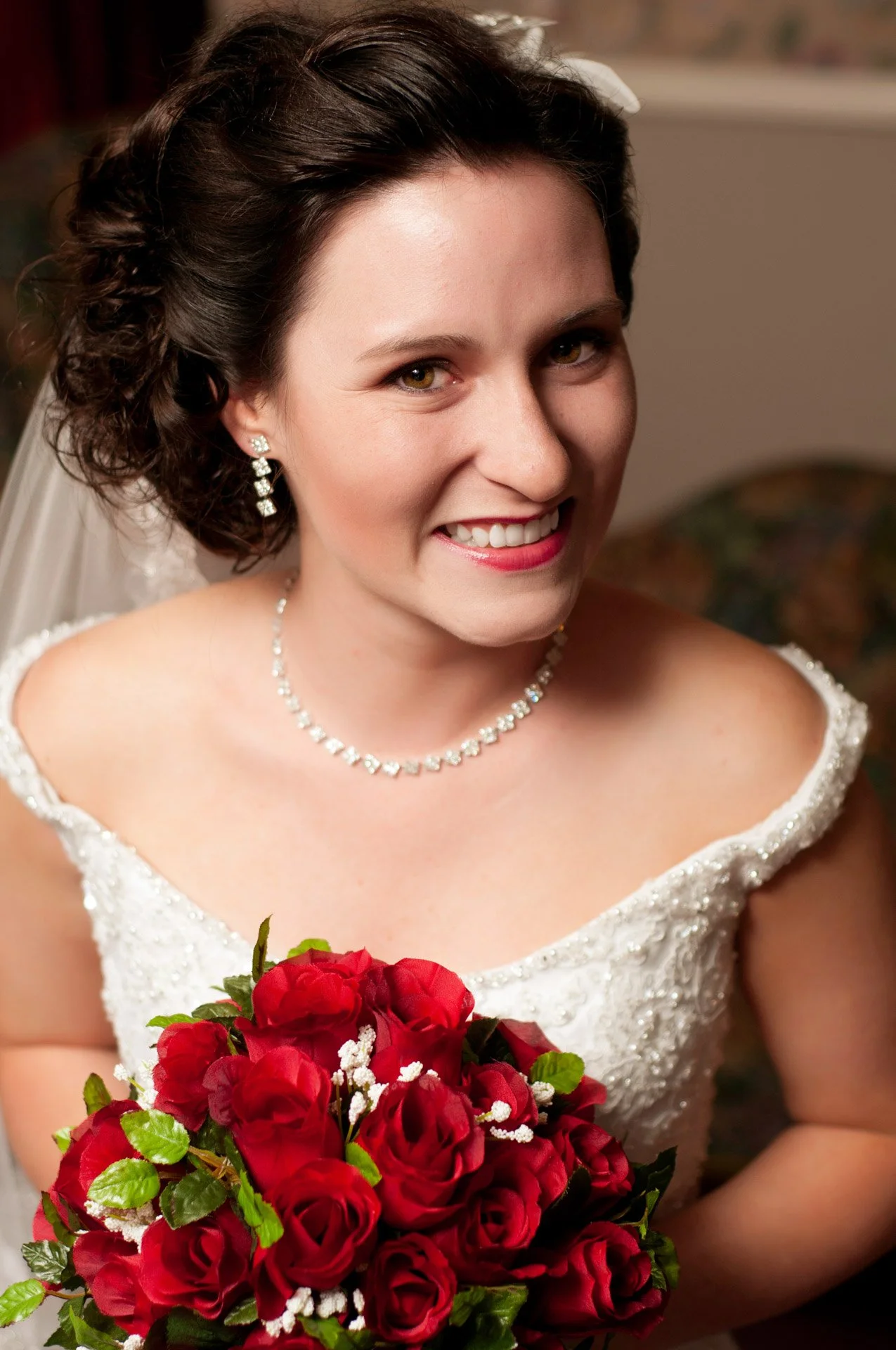 A smiling bride with dark brown curly hair, wearing a white wedding dress adorned with beads, diamond earrings, and a diamond necklace, holding a bouquet of red roses with green leaves and white baby's breath.