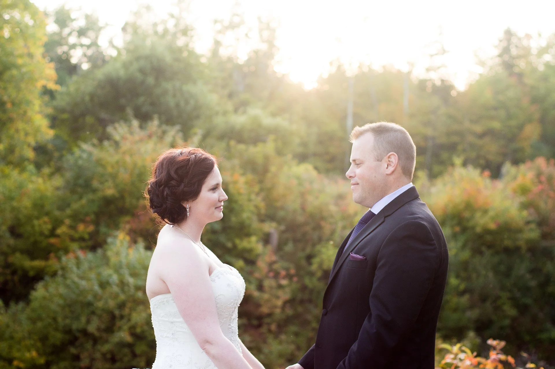 A bride and groom standing outdoors at sunset, holding hands and looking at each other with a backdrop of trees and autumn foliage.
