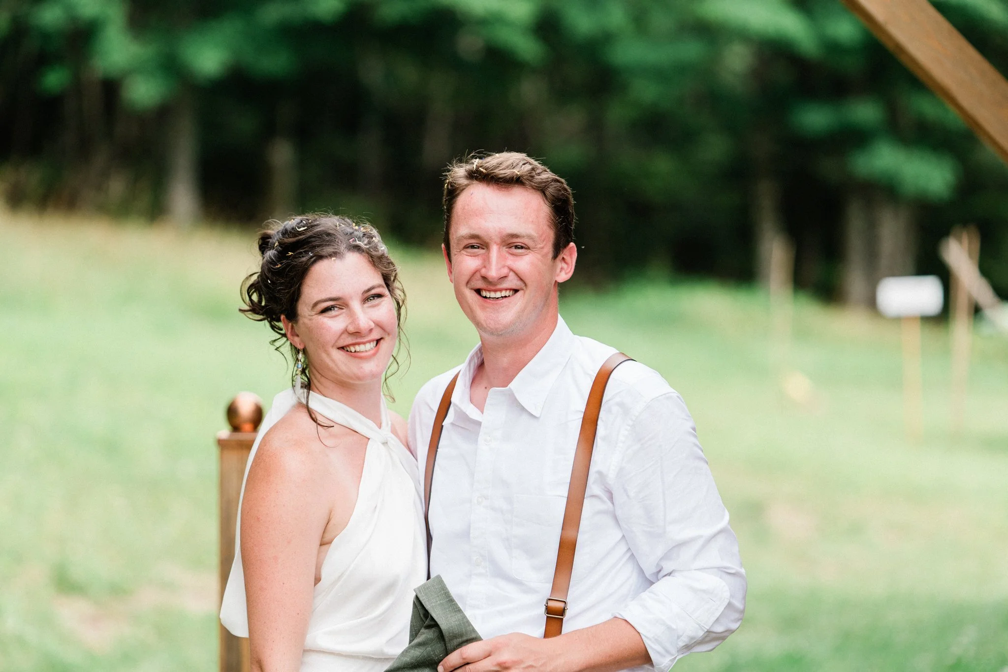 A smiling woman with curly hair and a white dress standing next to a smiling man with brown hair in a white shirt with suspenders, outdoors in a grassy area with trees in the background.