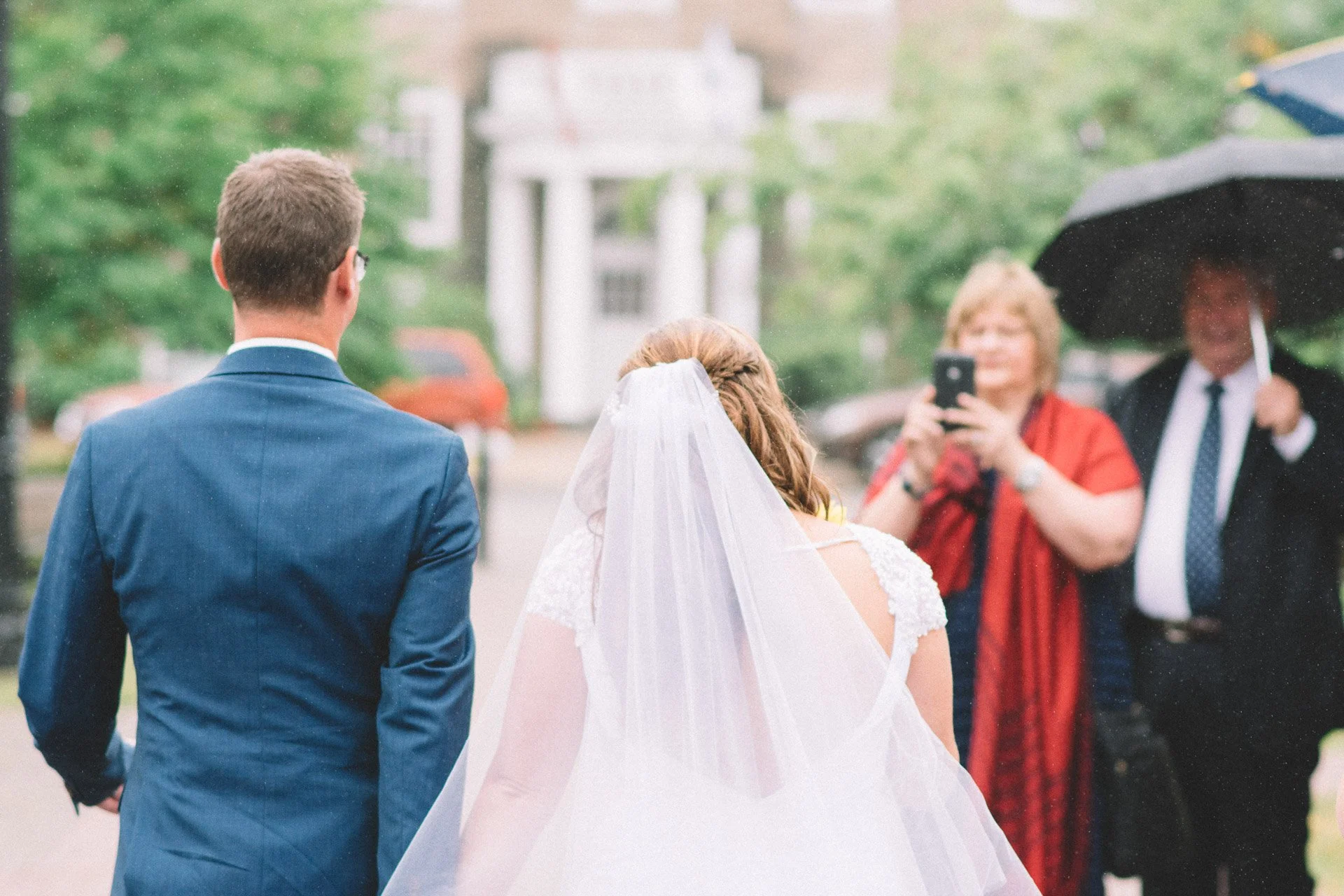 A bride and groom are seen from behind, standing outdoors on a rainy day, with women taking pictures of them under an umbrella.