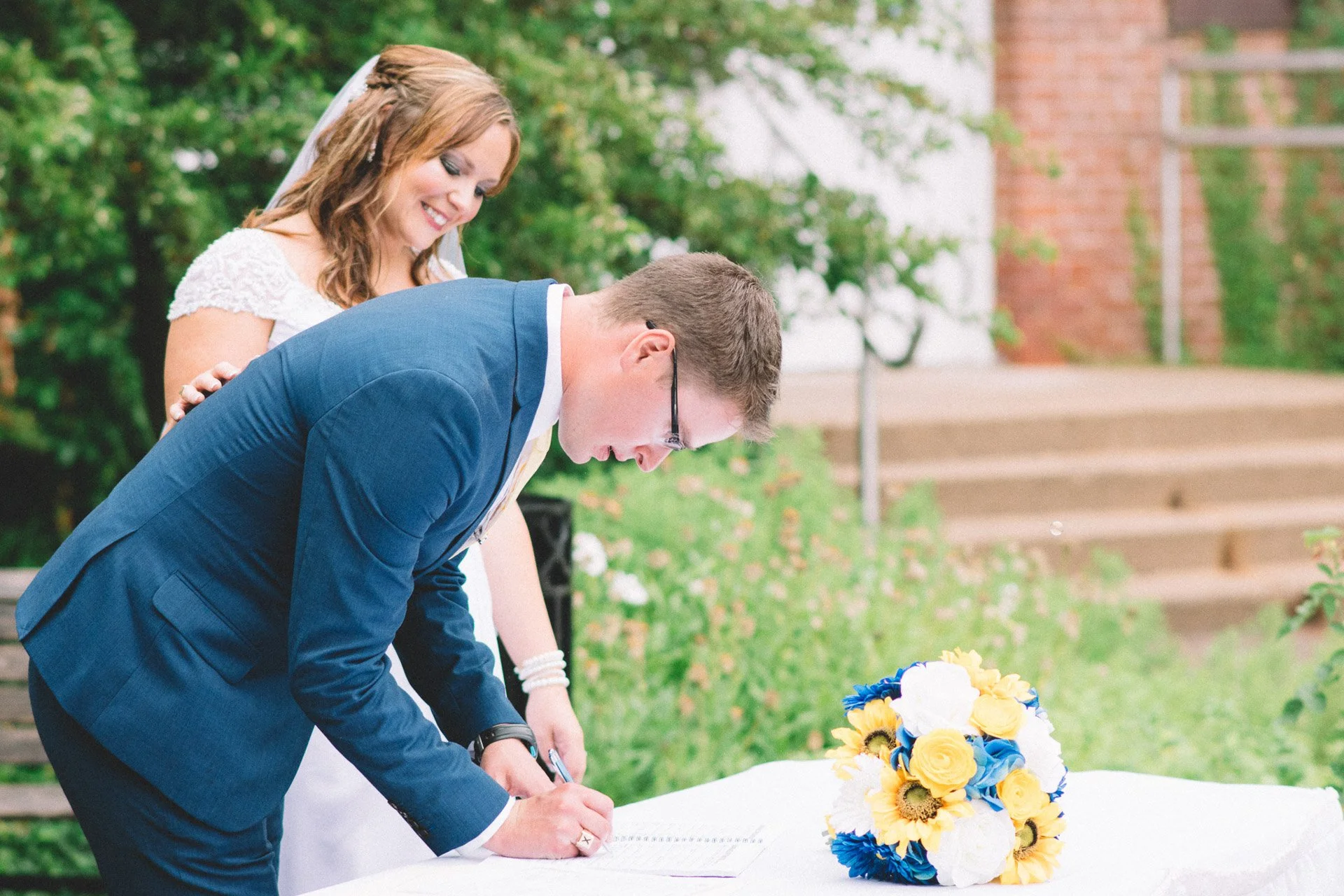 Bride and groom signing a marriage document outdoors, with a bouquet of yellow, white, and blue flowers on the table.