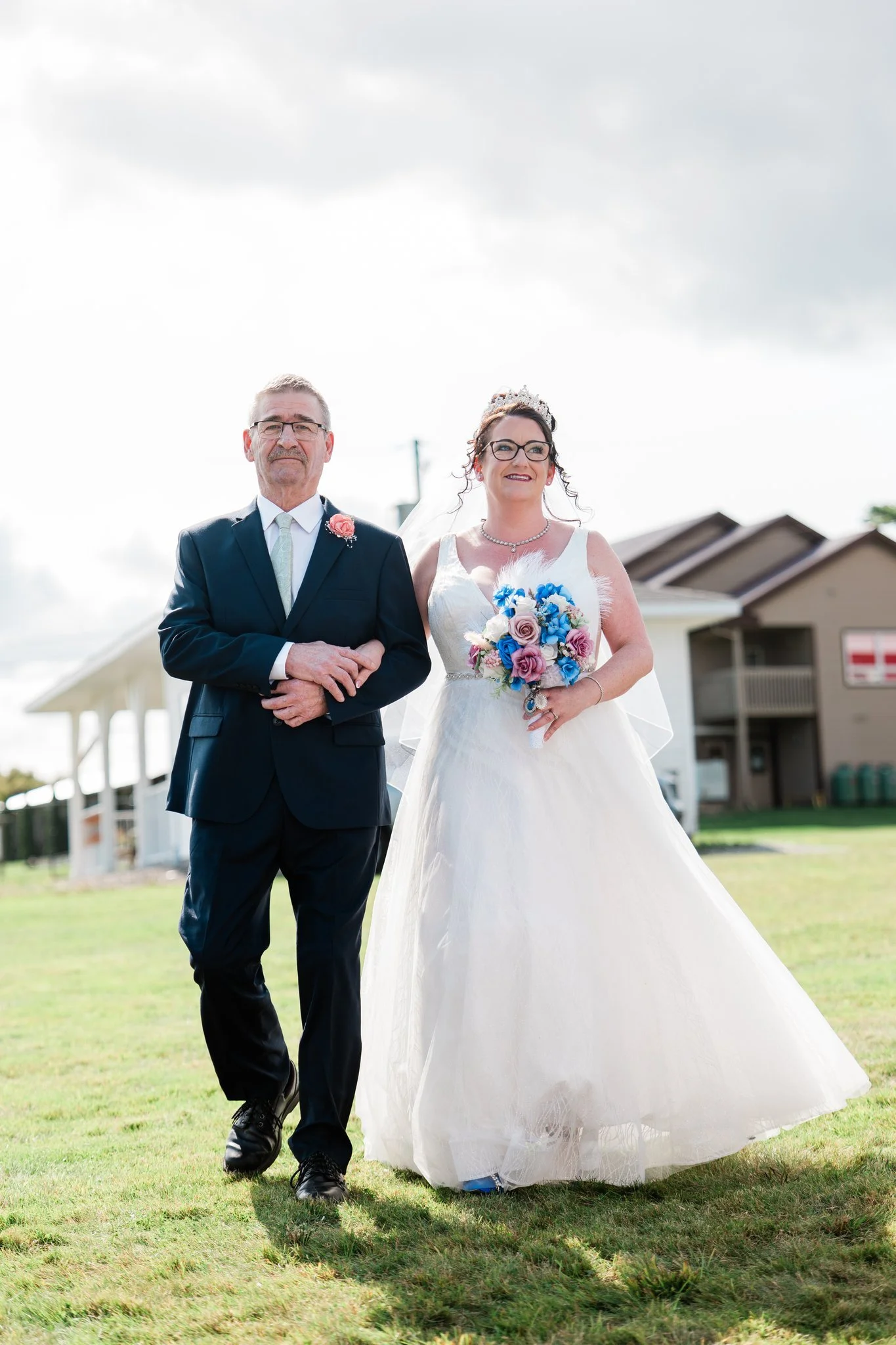 A bride in a white wedding dress holding a bouquet of blue and pink flowers walks alongside her father in a dark suit on a grassy area with a house-shaped building in the background.