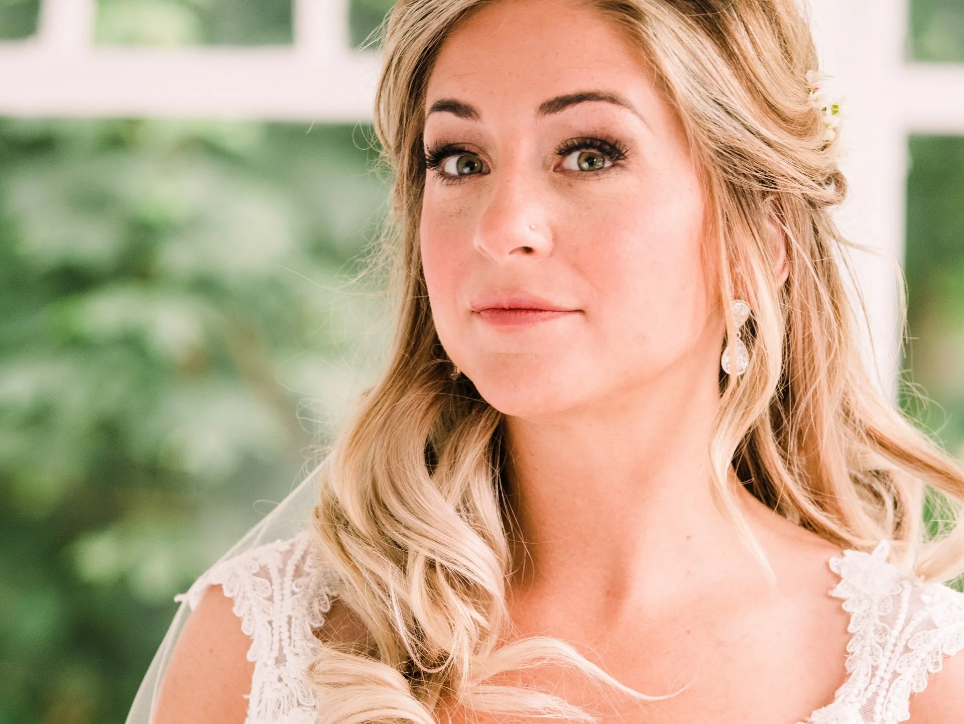 A bride with long blonde hair, wearing elegant earrings and a lace wedding dress, looking softly to the side with a blurred green background.