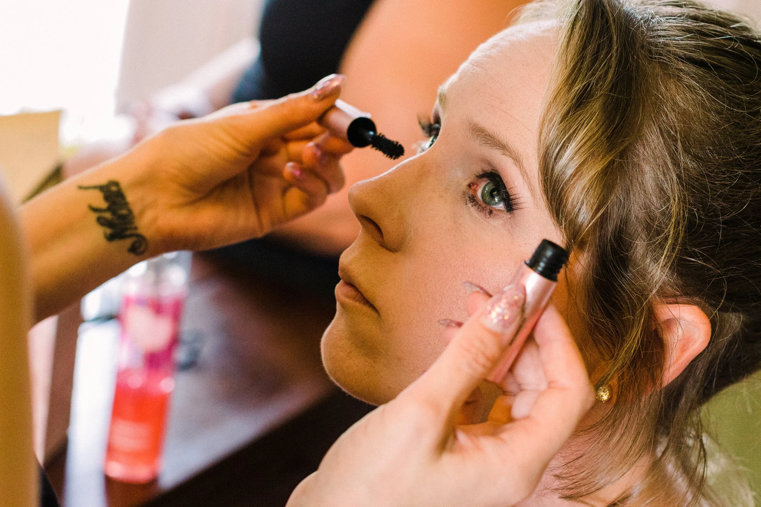 A makeup artist applies mascara to a young woman's eyelashes while she looks up. The woman has light skin, green eyes, and light brown hair styled in waves.