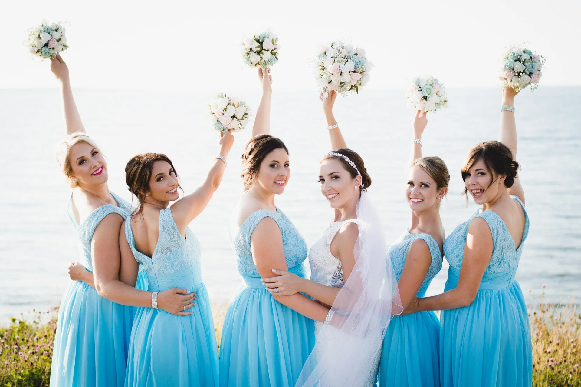 A bride and six bridesmaids at a beach, with the bridesmaids holding bouquets above their heads.