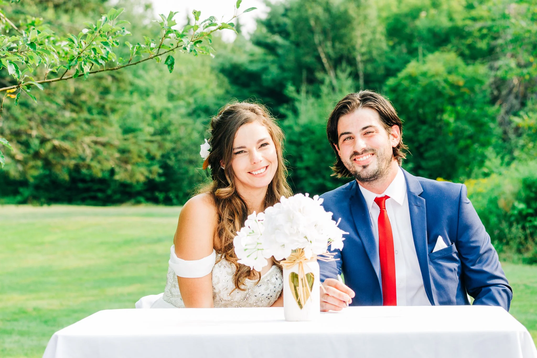 A bride and groom sitting at a table outdoors with a white wedding bouquet in front of them, surrounded by green trees and grass.