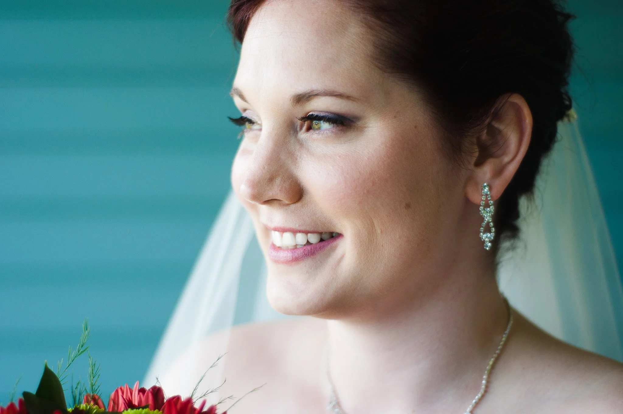 Close-up of a smiling bride with earrings and a necklace, holding a bouquet of red flowers, with a soft teal background.