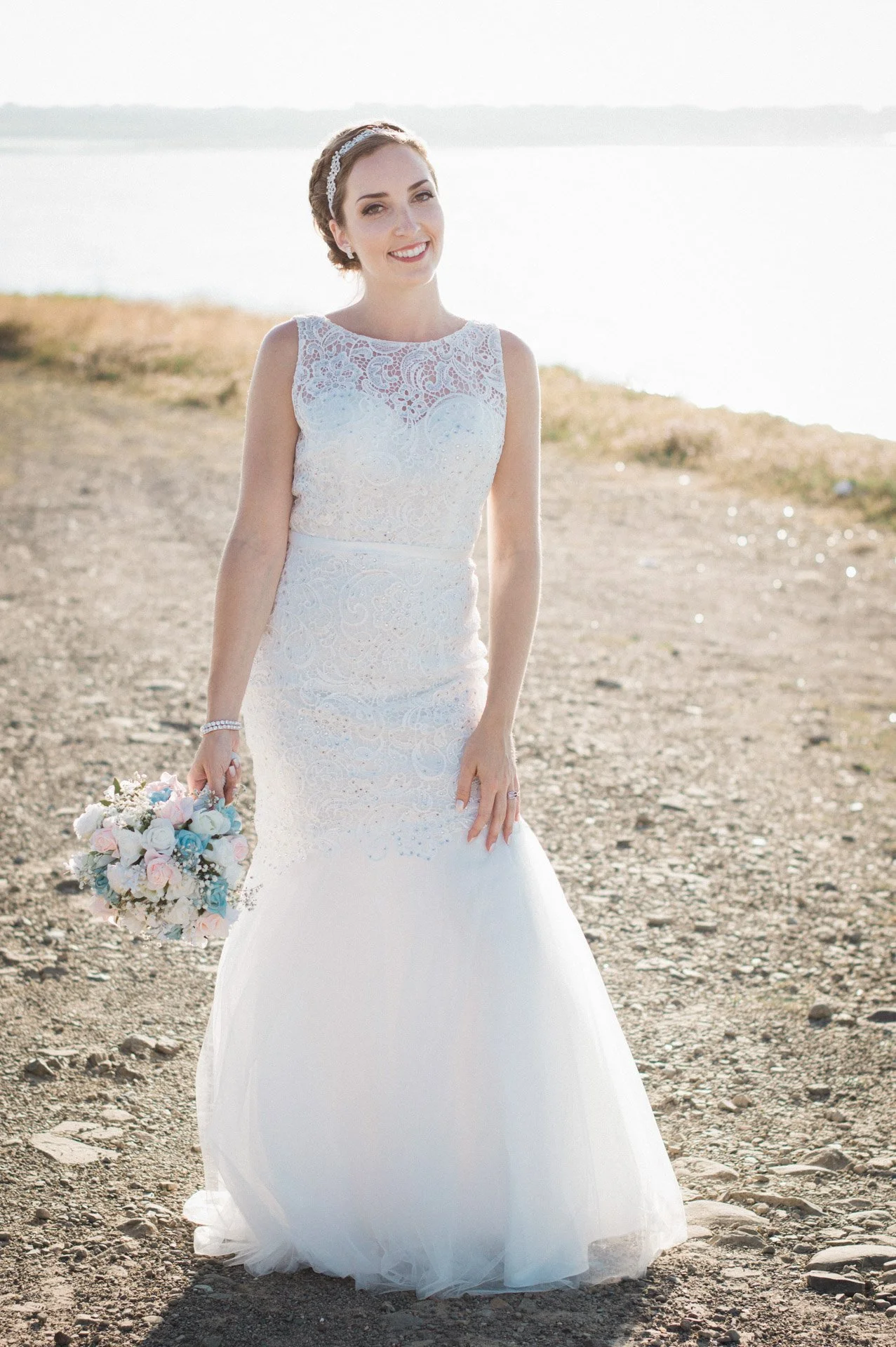 A bride in a white lace wedding gown holding a bouquet of pink and white roses on a rocky beach with water in the background.