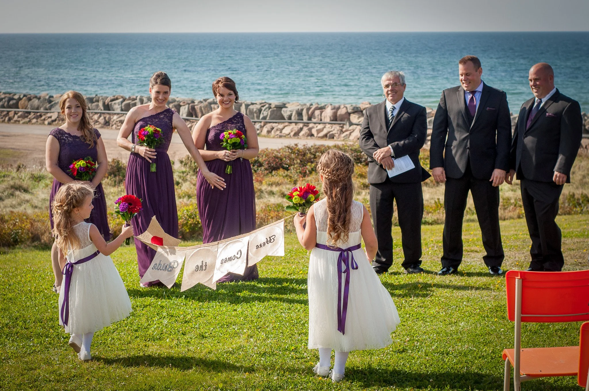 A wedding ceremony outdoors near the ocean with bridesmaids in purple dresses, flower girls in white dresses, and groomsmen in black suits, with a banner reading 'The Great Comes the Tea' and flower bouquets.