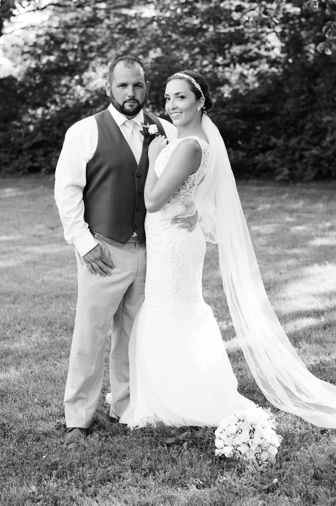 A bride and groom posing outdoors for a wedding photo, with trees in the background. The bride is smiling and wearing a lace wedding dress with a veil, while the groom is in a vest and dress shirt. A bouquet is on the ground near the bride.