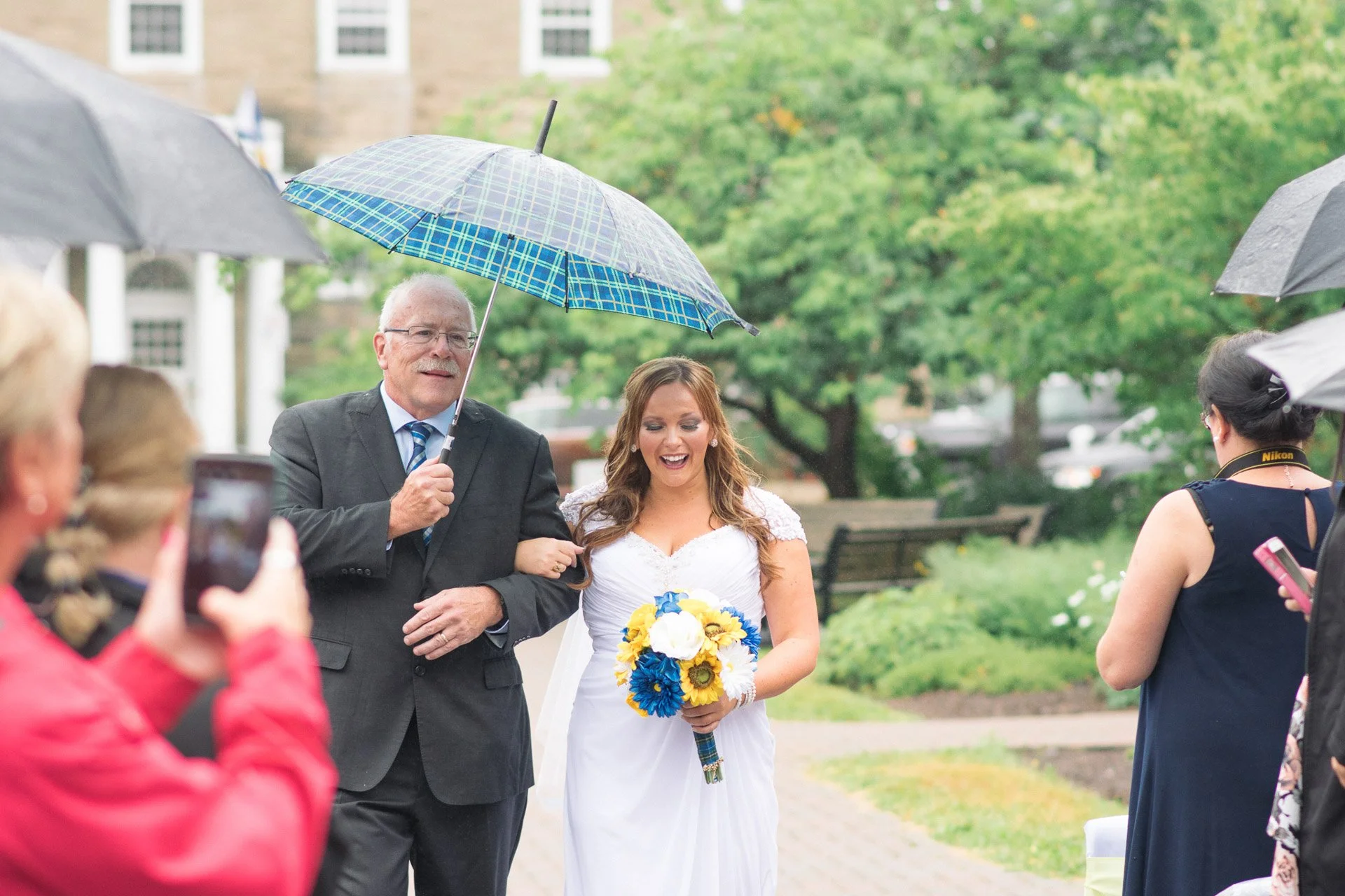 A bride in a white wedding dress holding a bouquet of yellow, blue, and white flowers, walking arm-in-arm with an older man who is holding an umbrella, while surrounded by guests with umbrellas outdoors in rainy weather.