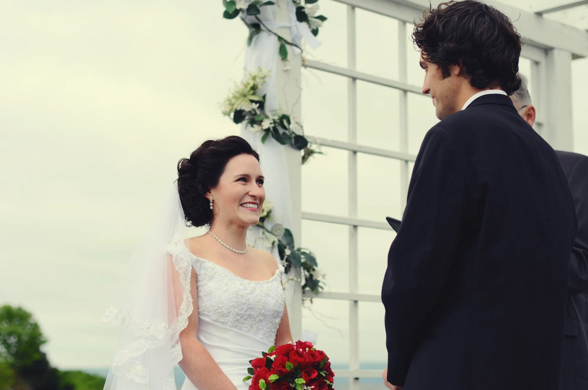 Bride and groom exchanging vows outdoors, with the bride holding a red bouquet, dressed in wedding attire, smiling at each other.