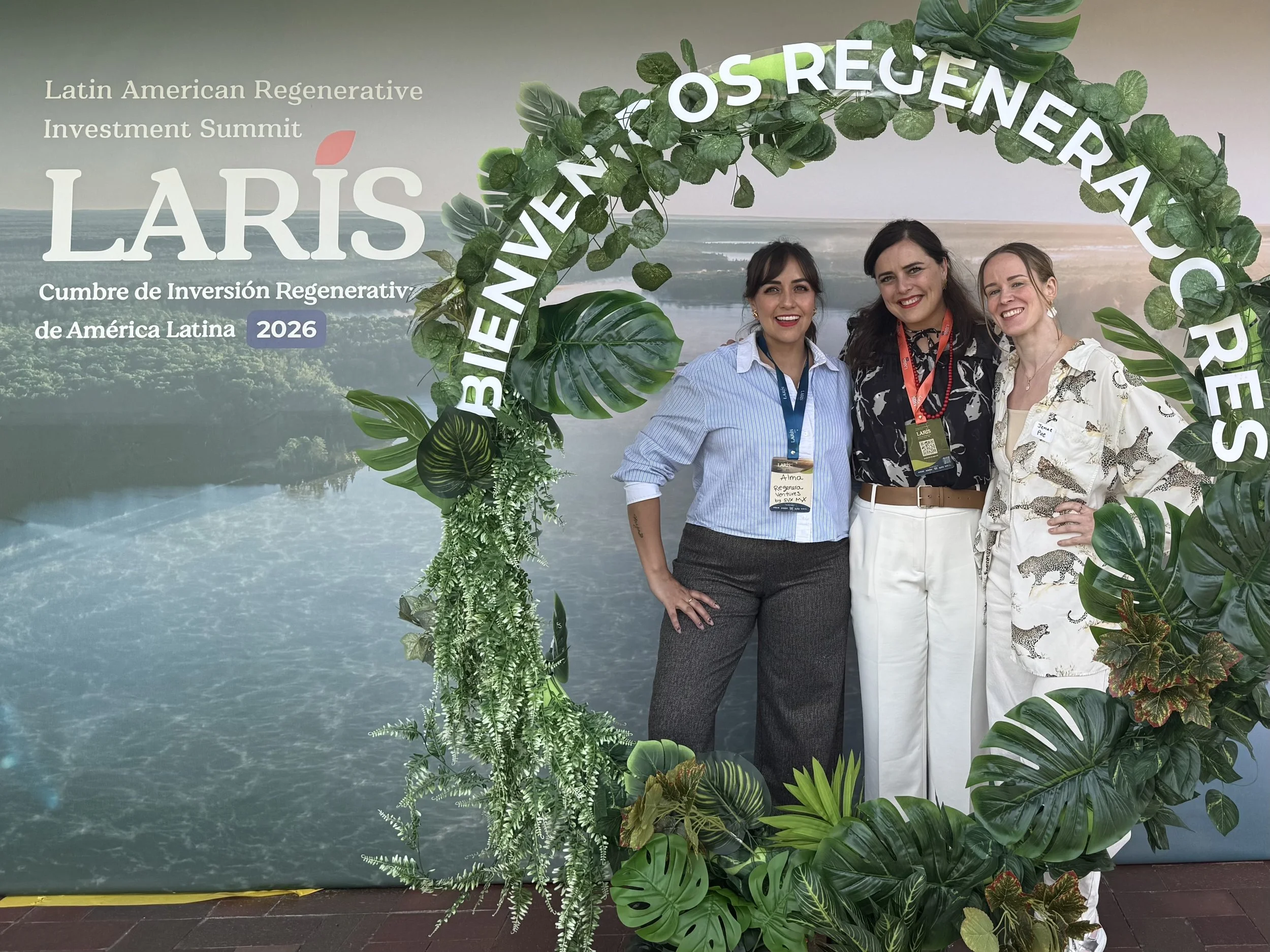 Three women standing together under a decorative arch made of green tropical leaves and plants at the Latin American Regenerative Investment Summit 2026.