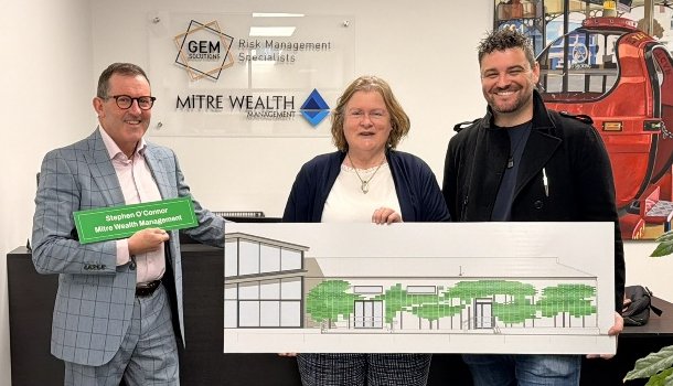 Three people standing indoors, holding a large architectural or building plan, with a wall behind them displaying logos for GEM Solutions, Risk Management Specialists, and MITRE WEALTH MANAGEMENT. The person on the left is holding a green nameplate, and there is a painting or poster of a red train in the background.