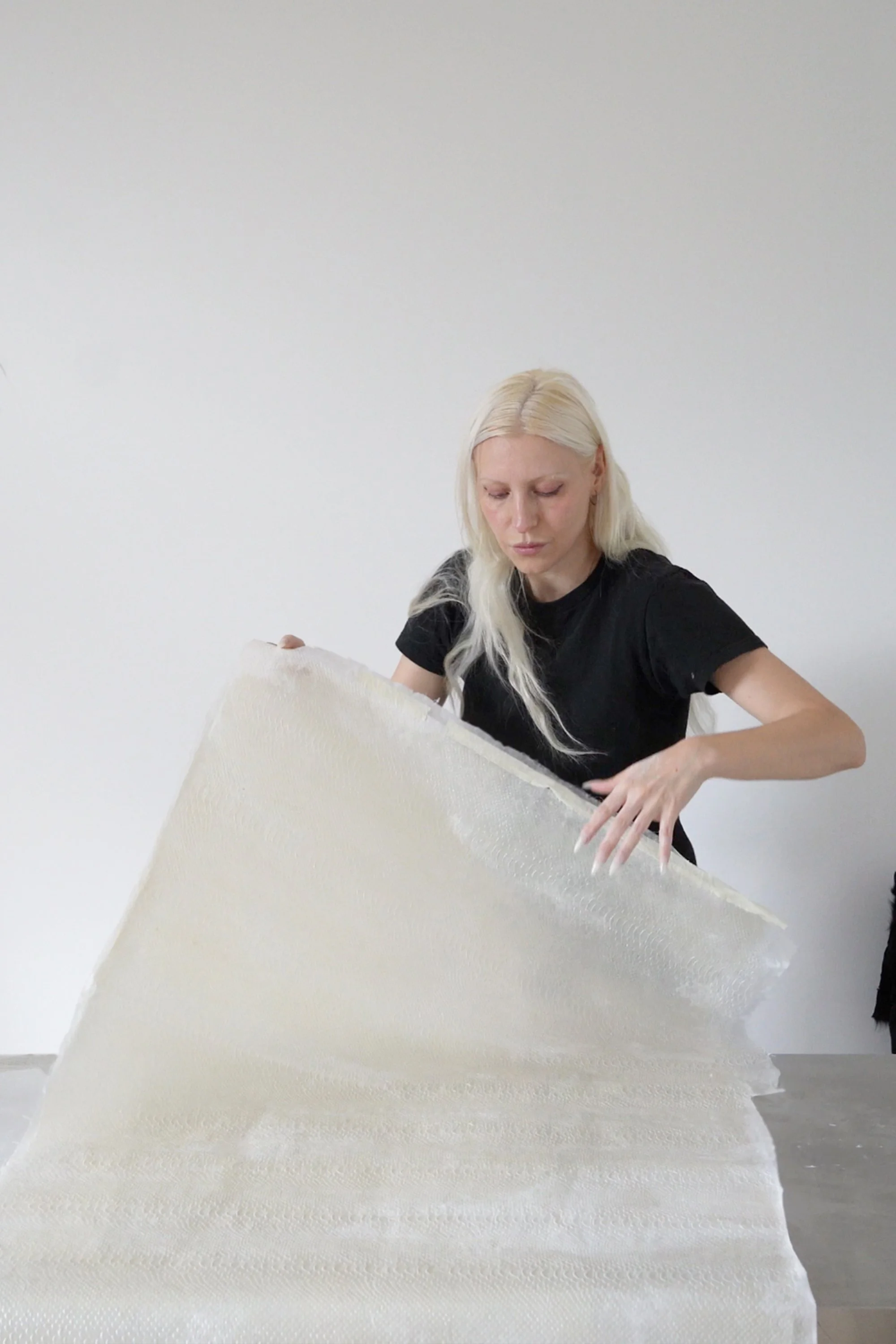 A woman with long blonde hair wearing a black shirt, holding a large white object, possibly a quilt or fabric, in front of a plain white wall.